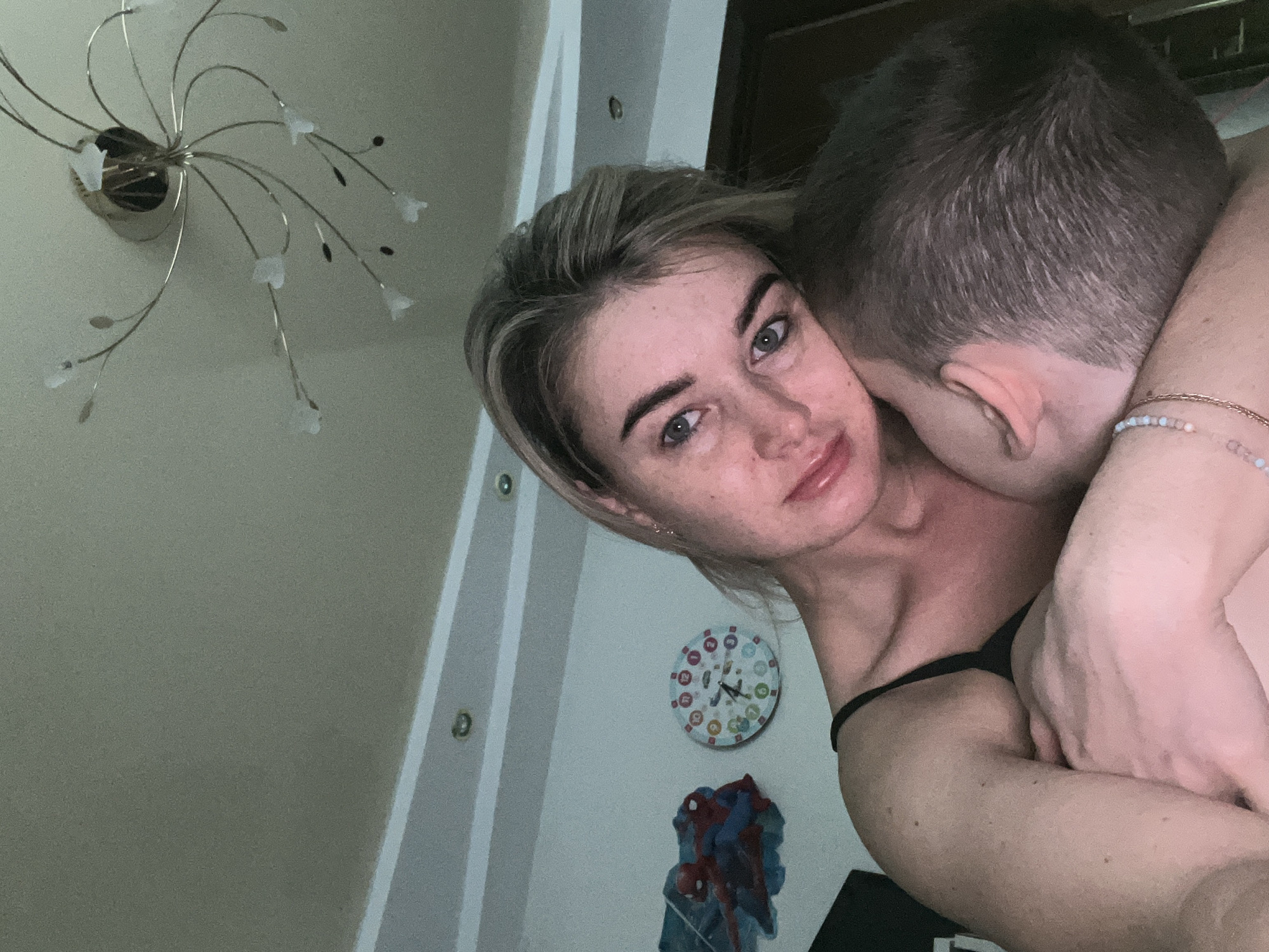 The image shows two people in an intimate embrace. The woman has short blonde hair and is looking at the camera. The man has short dark hair and is positioned behind her. A decorative chandelier is visible on the ceiling. On the wall, there is a small decorative clock and a picture of Spider-Man. The location is Chaikovsky, Russia.