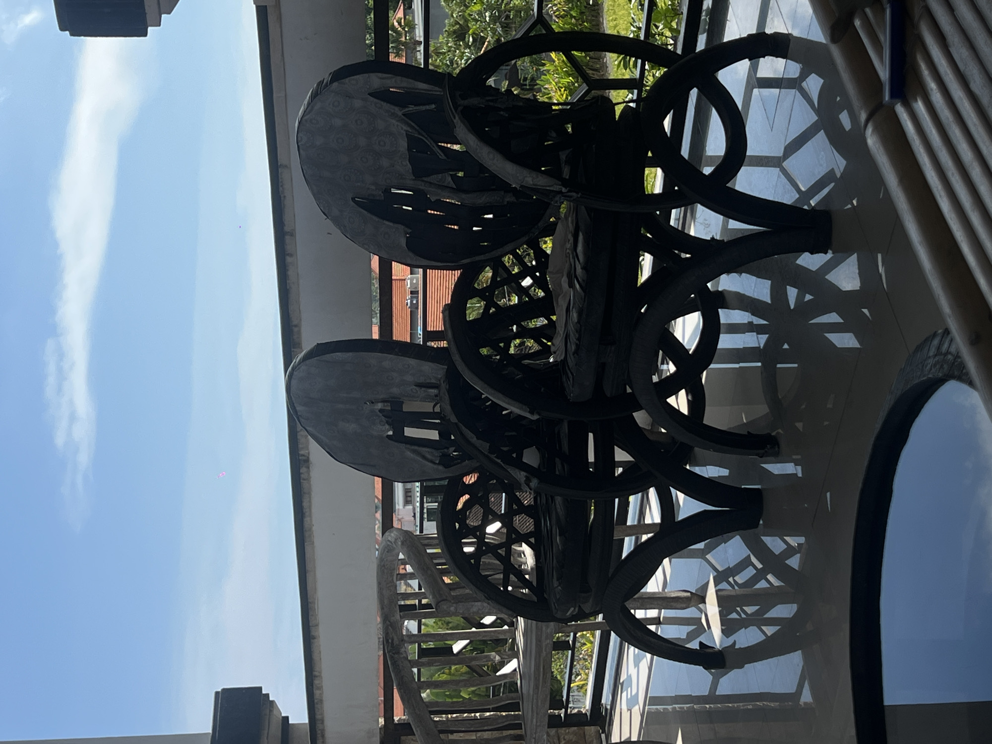 This image shows a set of three dark-colored, ornate wicker chairs placed on a glossy surface, likely a patio or balcony. The sky, visible in the background, indicates it is daytime. Based on the architectural style, foliage, and bright sky, the location is most likely Canggu, Indonesia. No people are visible. The objects are limited to the chairs and the surrounding environment.