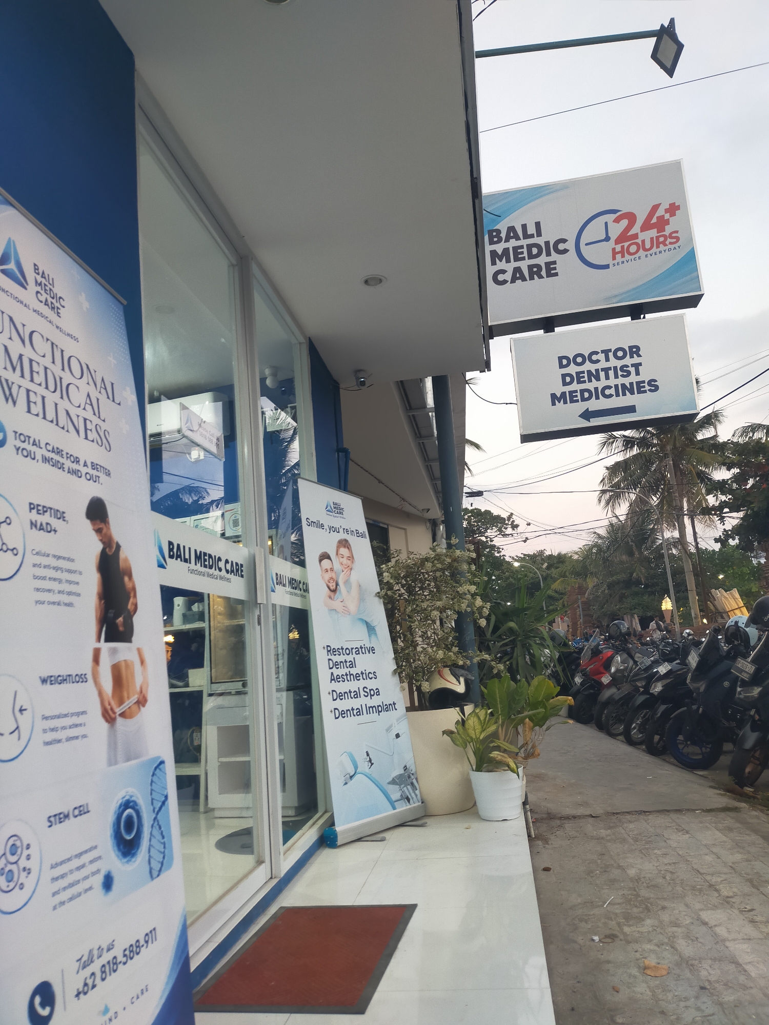 The image shows the exterior of "Bali Medic Care," a medical clinic in Kuta, Indonesia. The clinic features large glass windows and a blue and white color scheme on its signage and banners. One banner displays "FUNCTIONAL MEDICAL WELLNESS" with details on services like "PEPTIDE, NAD+" and "WEIGHTLOSS." Another banner features a couple smiling, with text highlighting "Restorative Dental Aesthetics," "Dental Spa," and "Dental Implant." Above the clinic entrance, two signs are visible: one reads "BALI MEDIC CARE 24 HOURS SERVICE EVERYDAY," and another beneath it states "DOCTOR DENTIST MEDICINES" with an arrow pointing left. The clinic is situated on a street with a row of parked motorcycles on the right side. The environment appears to be a commercial area with a sidewalk and a street, and palm trees are visible in the background. The sky is overcast, suggesting it is likely late afternoon or early morning. No people are clearly depicted as engaging in specific actions within the scene, though some figures are faintly visible through the clinic's windows.