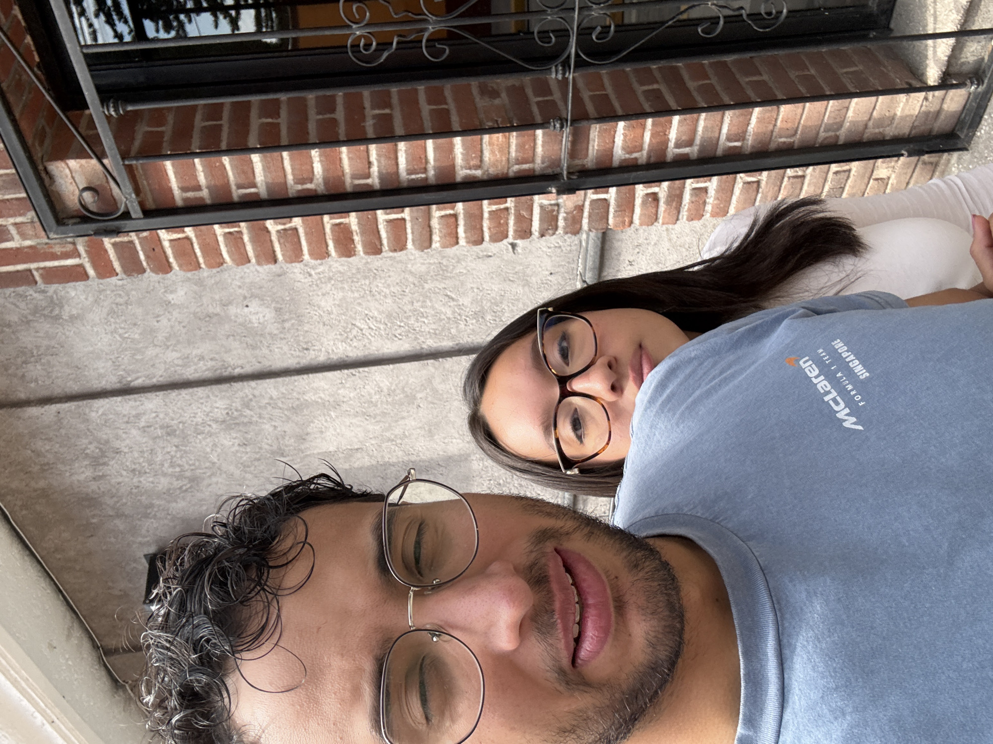 Two individuals are positioned closely together, implying a selfie or close proximity. The individual in the foreground is wearing glasses and has a beard. The person behind them also has glasses and dark hair. They are both wearing shirts. The background features a brick wall with a metal grate. The location is San Andrés Cholula, Mexico.