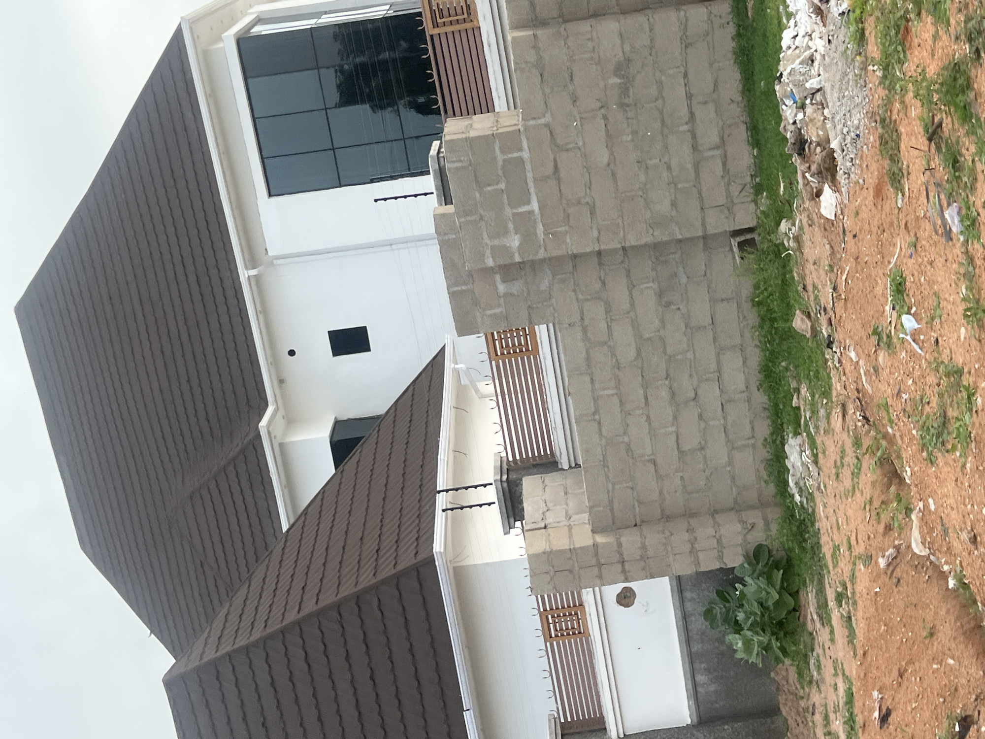 This media file captures a residential scene in Howayi, Nigeria, featuring two multi-story buildings, likely undergoing construction or recent renovation, next to an undeveloped plot of land. The image is shot from a slightly low angle, looking upwards towards the structures and the sky.
The prominent features are two modern-style buildings with dark brown corrugated roofs and white exterior walls. The closer building on the left shows two stories, with large, dark-tinted windows on the upper floor and a smaller dark window on the lower floor. It has a balcony with a brown vertical slatted railing. In front of this building, a wall made of grey concrete blocks is partially built, indicating ongoing construction or a boundary.
The second building, visible behind and to the right of the first, shares a similar architectural style with a brown corrugated roof, white walls, and a balcony featuring a matching brown slatted railing. A smaller brown wooden structure is visible on one of its lower balconies.
No people are visible in the image, suggesting a quiet moment at a construction site or residential area. The ground area to the right consists of reddish-brown dirt, characteristic of the region, mixed with sparse green grass and scattered debris such as plastic and paper, suggesting undeveloped or neglected land adjacent to the buildings. A patch of vibrant green foliage grows near the base of the concrete block wall, close to a white door or panel.
The sky is overcast, indicating daytime conditions without direct sunlight. There is no visible text in the image.