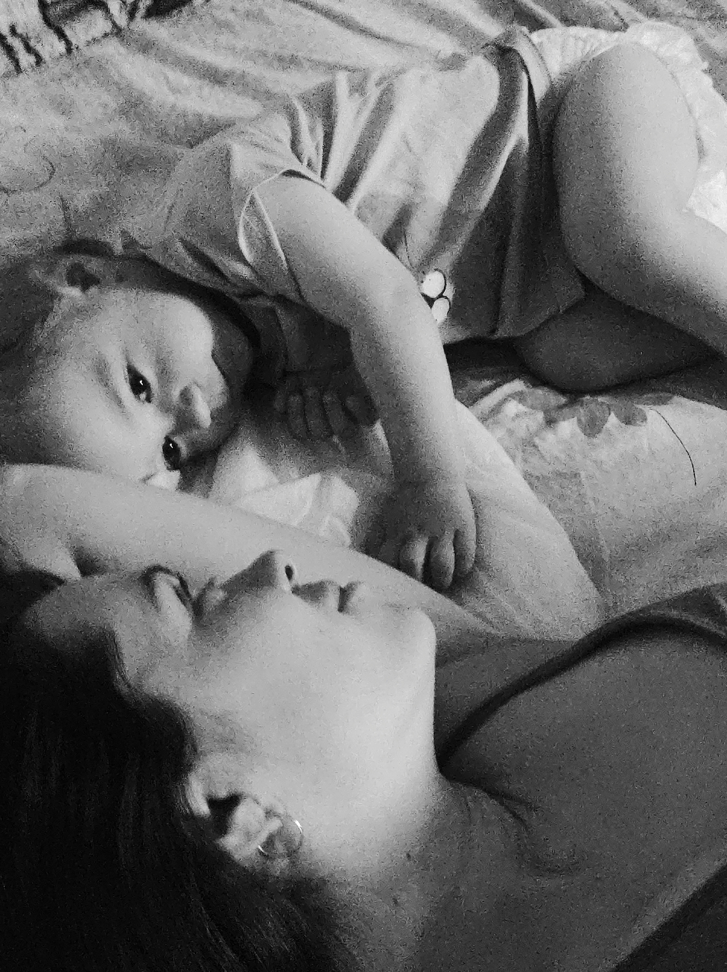 A black and white photograph shows a woman lying on her back with a baby positioned on top of her chest. The woman's head is turned to the side, her eyes closed. The baby is lying partially on their side, their eyes open and looking at the camera. Both appear to be lying on a bed covered with a patterned sheet. The location is unknown.