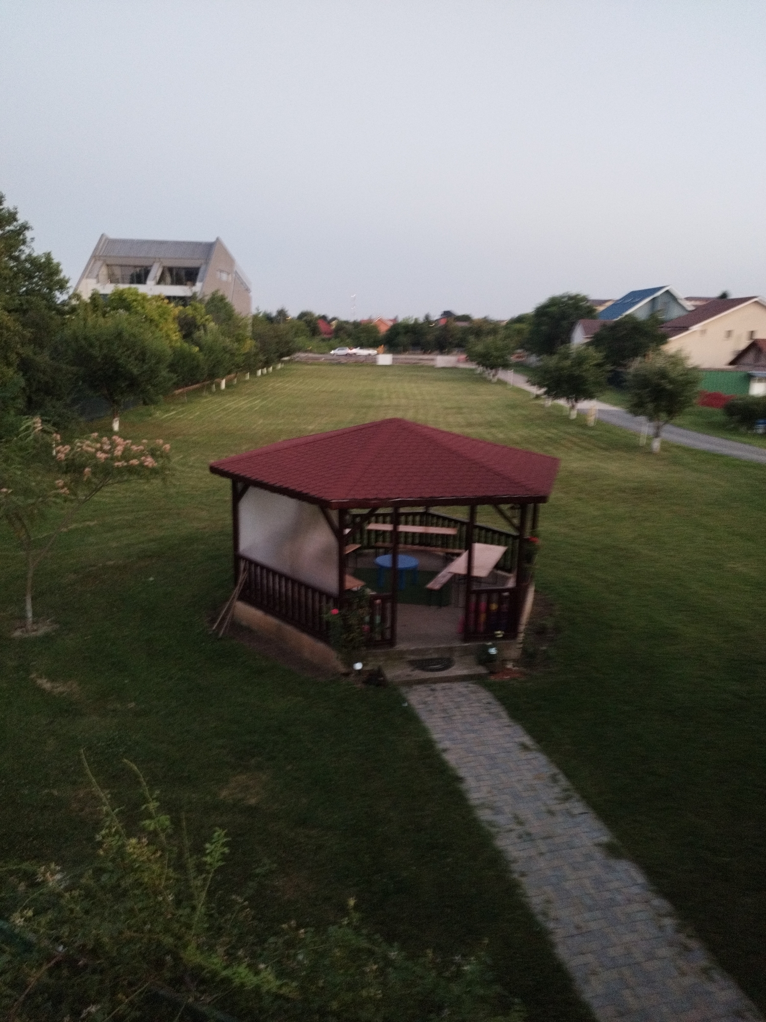 The image depicts a backyard scene with a red-roofed gazebo in the foreground. The gazebo has white siding and is furnished with seating and a blue table. A paved pathway leads to the gazebo through a well-maintained lawn. In the background, several houses with pitched roofs are visible, along with trees and a street. The sky is overcast and light. The scene appears to be a suburban residential area, possibly in the city of Bucharest, Romania, given the architectural style of the houses and the overall landscaping. There are no people visible in the image, and no activities are taking place. The image captures a quiet, late afternoon or early evening atmosphere.