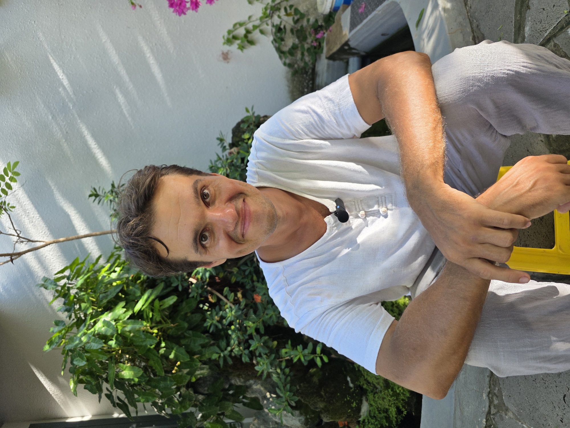 A man with short, dark hair and a fair complexion is sitting outside in Mũi Né, Vietnam. He is wearing a white, short-sleeved shirt with buttons and light grey pants. He is seated on a yellow object, likely a stool or part of a larger structure. His arms are crossed, and he is looking directly at the camera with a slight smile. Behind him is a white wall partially covered by lush green foliage and a pink flowering plant above. The environment is bright, with natural light casting shadows on the wall.