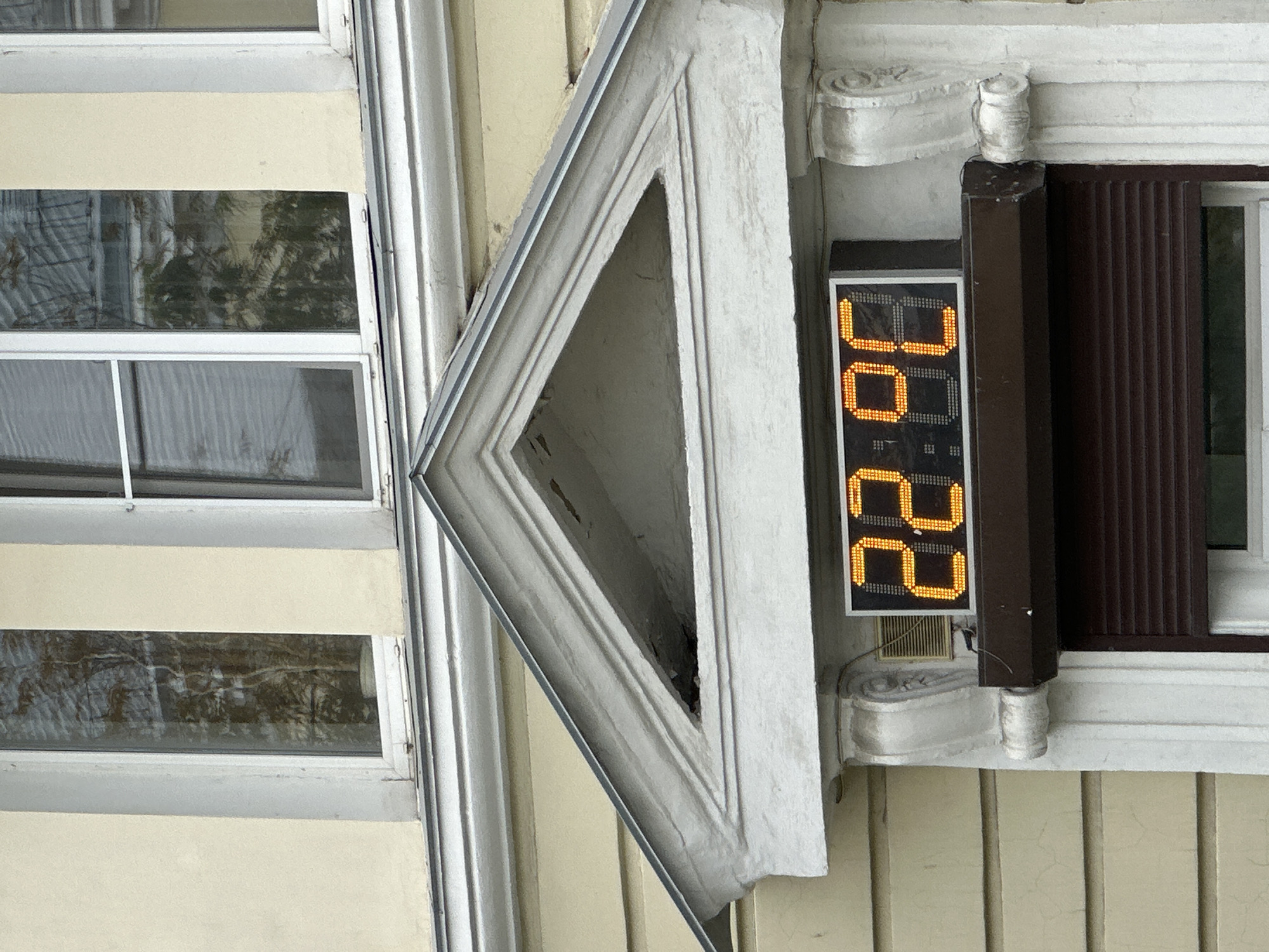 An outdoor digital clock displays "22.98" in orange digits, mounted above a closed brown rolling shutter. The clock is situated on a cream-colored building with white decorative trim. Above the clock, a triangular pediment with ornate plasterwork is visible. In the background, two windows with white frames show reflections of trees. The image is an eye-level, medium shot focused on the clock and its immediate surroundings.