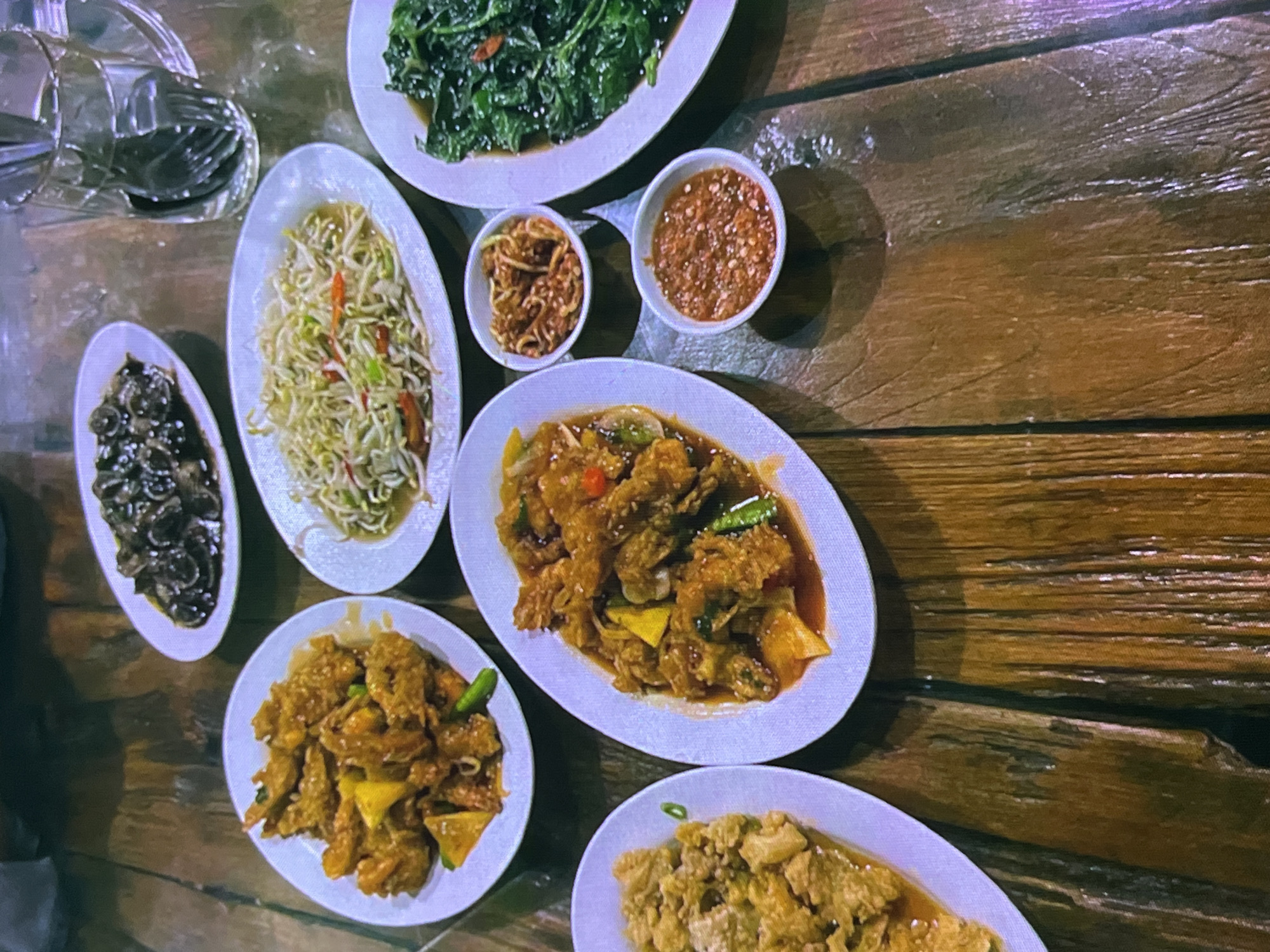 The image depicts a table laden with several dishes of Indonesian food, set on a rustic wooden surface. The food appears to be served in white oval platters and small white bowls.
In the foreground and center, a white oval platter contains fried food coated in a reddish-orange sauce, possibly sweet and sour or a chili-based sauce, with visible pieces of green chili and what looks like pineapple chunks. To the left of this, another white oval platter holds a dish of golden-brown, bite-sized fried pieces, likely chicken or seafood, seasoned with herbs.
Slightly further back and to the right, a large white oval platter is filled with a generous portion of stir-fried leafy green vegetables, possibly water spinach or kale, seasoned with chili. Next to this, a smaller white oval platter contains a light-colored stir-fry of bean sprouts and thin strips of vegetables, including red chili.
In front of the bean sprout dish, two small white bowls are visible. The top bowl contains a dark reddish-brown condiment, possibly fried shallots mixed with chili. The bottom bowl holds a vibrant red, chunky chili sauce.
In the upper portion of the image, a white oval platter holds a dark, glistening dish, appearing to be mushrooms or some sort of dark seafood in a glossy sauce. To the right of this, a clear glass pitcher filled with water and dark utensils is visible.
The overall setting is casual, suggesting a restaurant or home dining environment. The wooden table has a distressed, polished finish, with visible grain and some water marks. No people are clearly visible in the frame, though a person's arm or shoulder might be suggested in the upper left corner, out of focus. The lighting is somewhat dim, casting a warm glow over the food. The scene is located in Cadek, Indonesia.