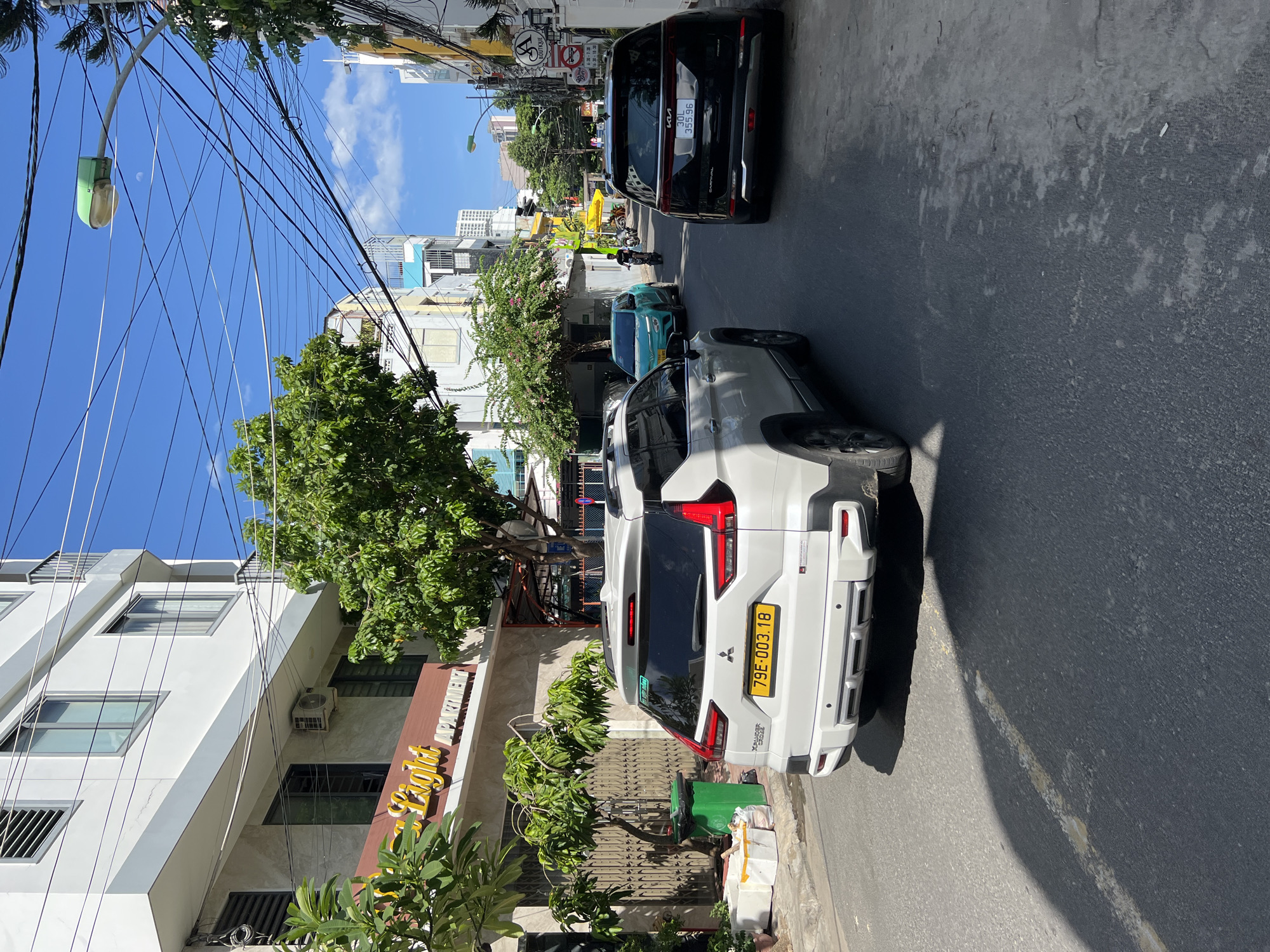 This image depicts a street scene in Nha Trang, Vietnam, under a clear blue sky with scattered clouds. The foreground is dominated by the rear of a white Mitsubishi Xpander Cross SUV, displaying a Vietnamese license plate "79E-003.18". To its right, a black Kia Carnival SUV is parked, with a Vietnamese license plate "30L 355.96" visible.
The street is paved and appears to be in an urban setting, lined with buildings of varying heights and architectural styles. On the left, a modern white building with multiple windows and balconies features signage reading "Light APARTMENT". Adjacent to it is a green trash bin. Further down the street, a turquoise car is parked.
Overhead, a complex network of electrical wires crisscrosses the sky, supported by poles, one of which holds a green street lamp. Trees with lush green foliage are visible along the street, adding to the urban greenery. The overall atmosphere suggests a typical day in a Vietnamese city, with vehicles, buildings, and infrastructure coexisting.