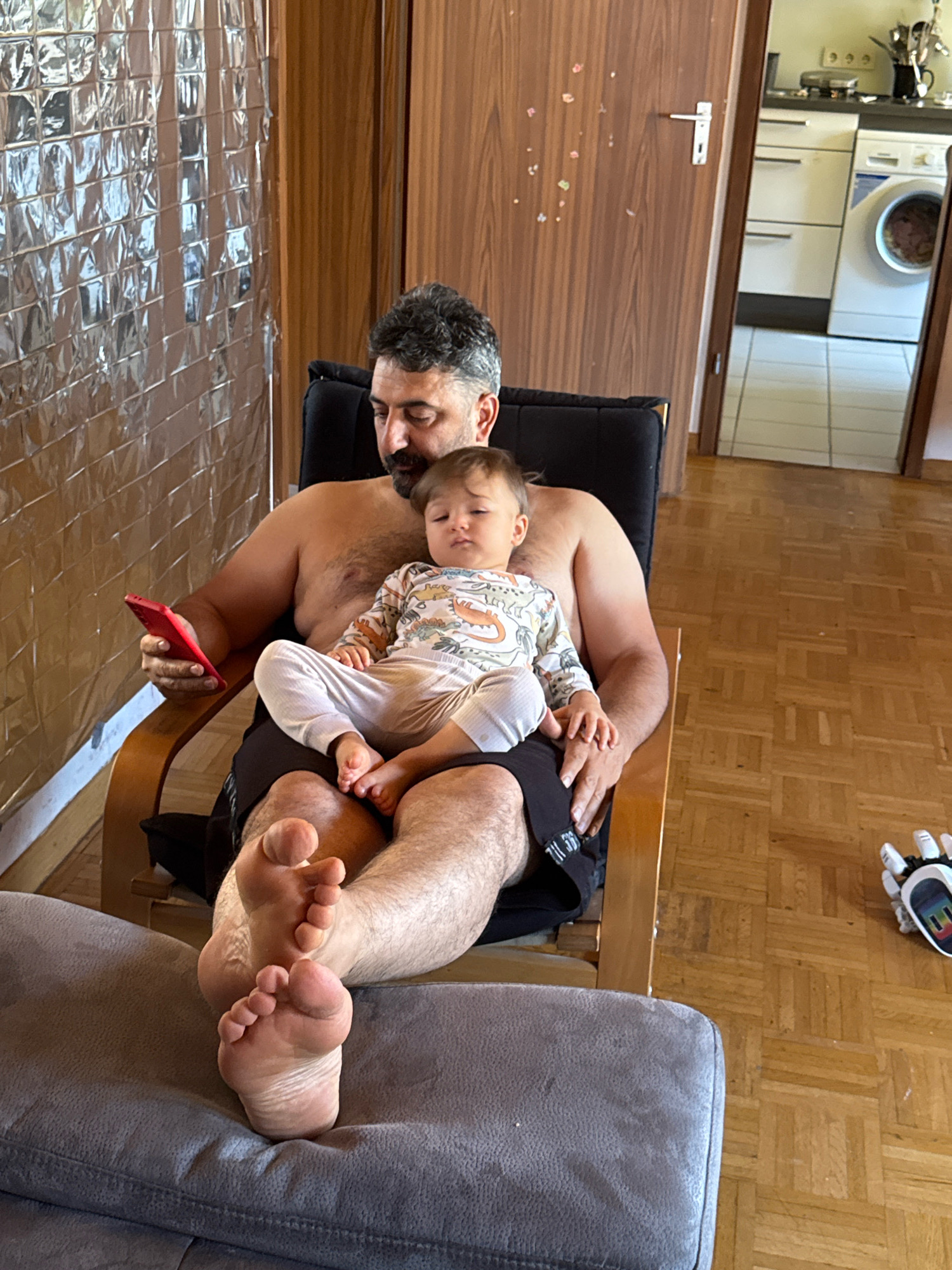 In Überherrn, Germany, a shirtless man with a beard sits in a black chair, holding a red mobile phone in his left hand. A baby, wearing a light-colored onesie with an animal print, sits on his lap. The baby's hands rest on the man's leg. The man's legs are extended, with his feet visible. The background shows a wall with a textured, reflective surface, a wooden door, and a partially visible kitchen area with a washing machine. A grey sofa is in the foreground.
