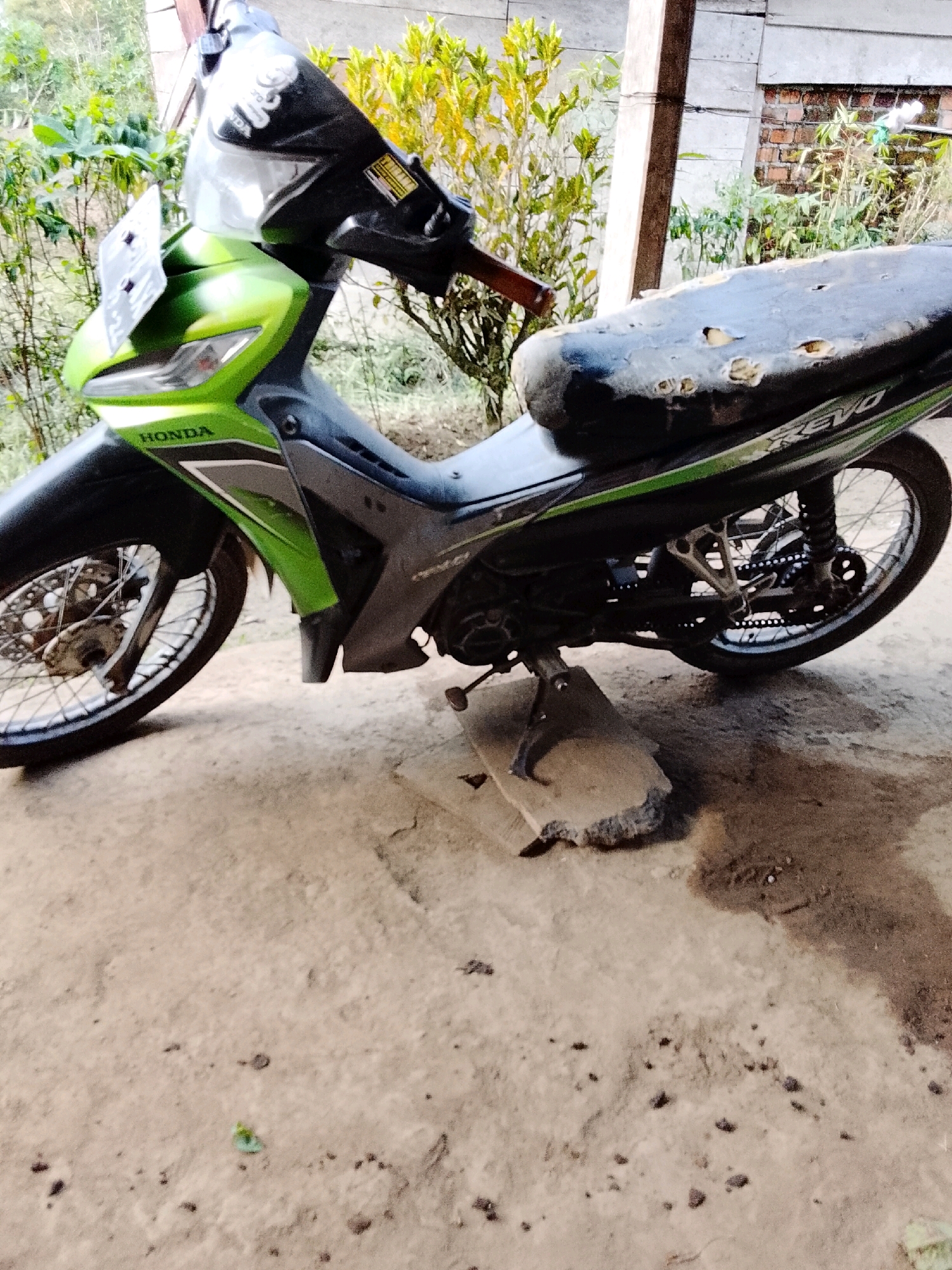 A green and gray Honda Revo motorcycle is parked on a dirt surface. The motorcycle's seat is ripped and appears worn. The front of the motorcycle features a green fairing and a license plate. A piece of concrete is placed under the motorcycle's kickstand, suggesting it is propped up. In the background, there are plants and a brick wall, indicating an outdoor setting. The scene appears to be in a residential area.