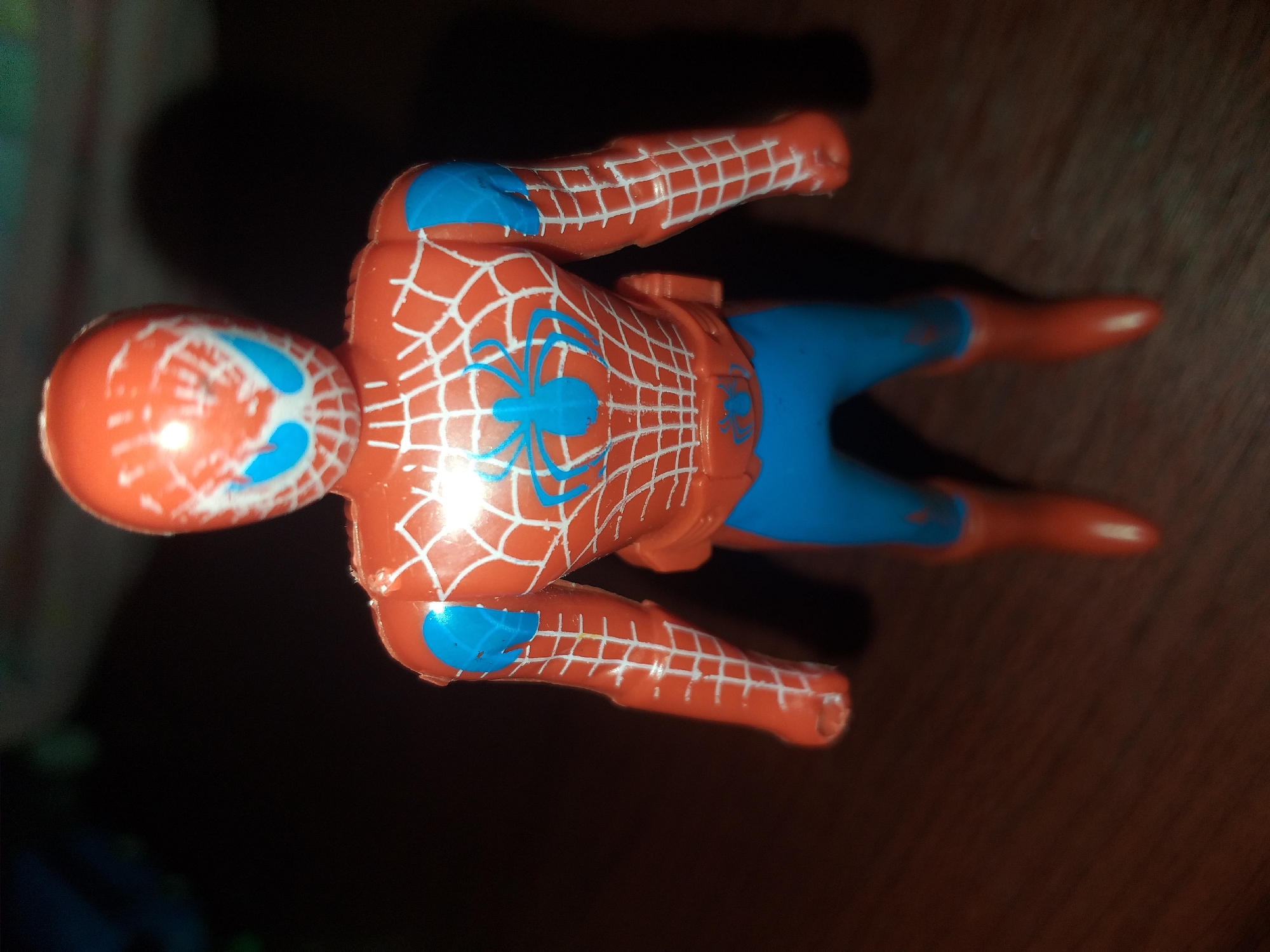 A Spiderman action figure is shown standing upright on a wooden surface. The figure is predominantly red and blue, with white webbing details across its body. It has blue eye lenses on its mask and a blue spider emblem on its chest. The figure's right arm and hand are visible. The background is dark and out of focus, with some hints of green and blue objects in the upper left corner. The lighting suggests a dim indoor environment. No people are present, and the action figure is the sole subject. The location is unknown, and there is no indication of Air Jamban, Indonesia.