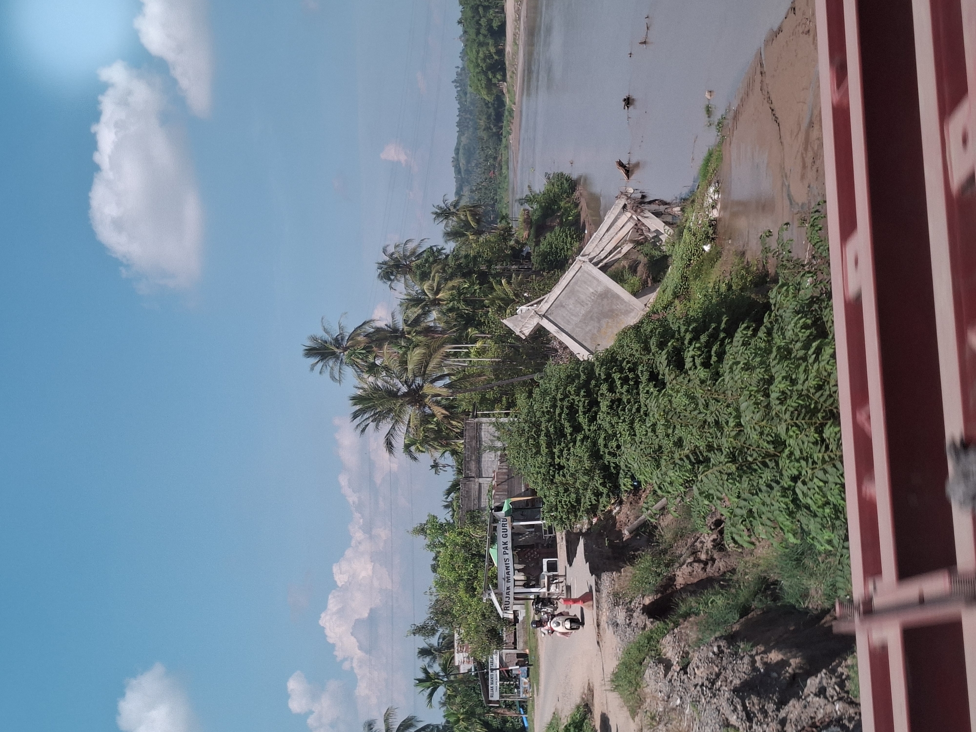 The image shows a rural scene in Blang Mee, Indonesia, featuring a wide river under a bright, partially cloudy sky. On the left bank of the river, a road curves past several small buildings, including what appears to be a food stall with a sign reading "RUJIAK MANIS PAK GURU". A person is riding a scooter past this stall. Adjacent to the road, a section of the embankment has collapsed, revealing exposed soil and debris, with lush green vegetation growing on the remaining slopes.
Further along the riverbank, a large concrete structure, possibly a bridge abutment or a section of a building, has collapsed and is partially submerged in the water. Debris from the collapse is scattered around it. The river itself appears to be muddy, with some logs and other natural debris floating on its surface. The background on the right side of the image shows a densely forested landscape with rolling hills. In the foreground, a red railing suggests the image was taken from a bridge or elevated viewpoint. The overall impression is one of a tropical, somewhat undeveloped environment, with signs of recent damage or disrepair.