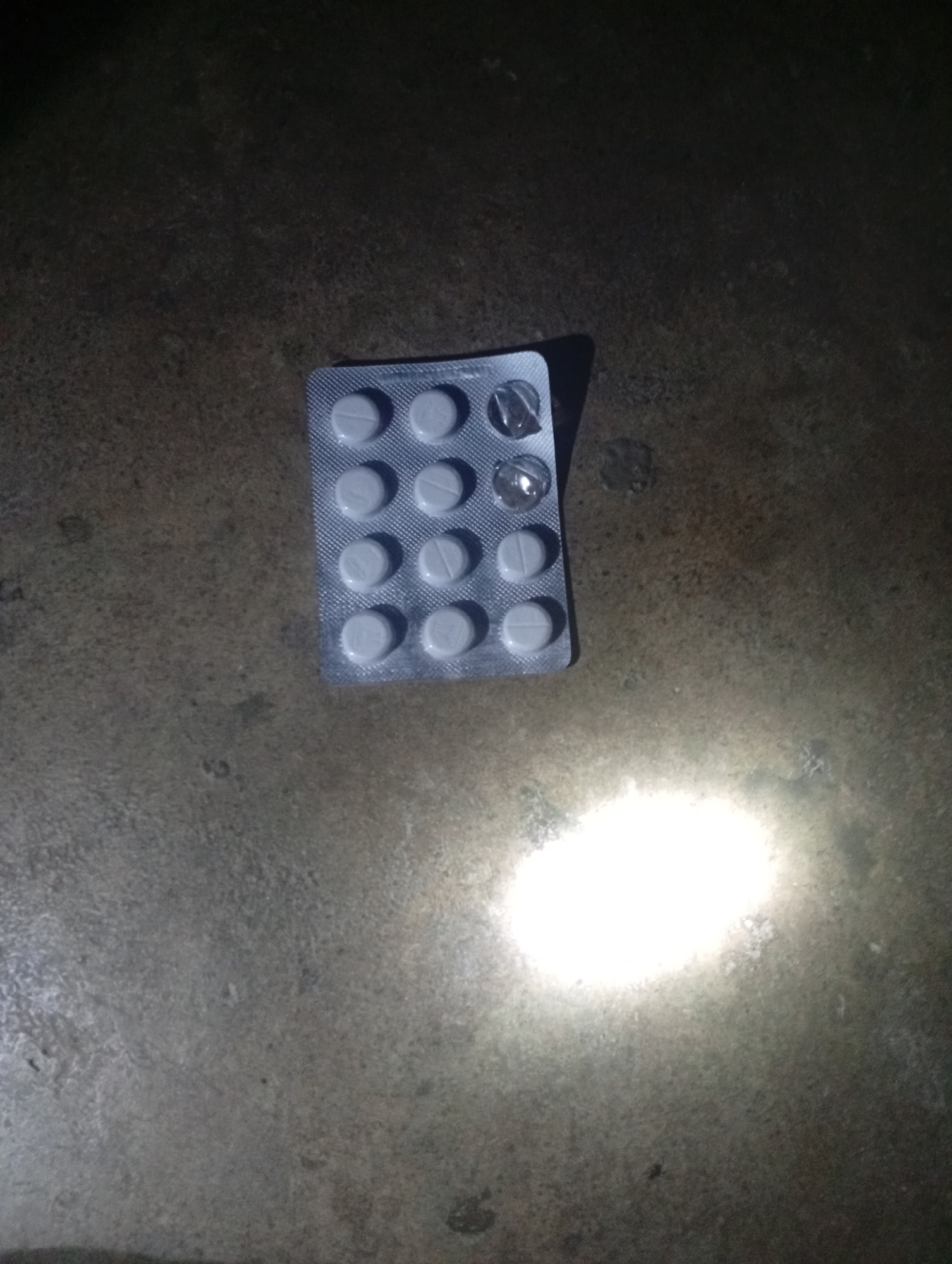 A partially used blister pack of white, round pills lies on a dark, textured surface that appears to be concrete. The blister pack is arranged in a grid of four rows and three columns, with some pills missing. A bright light source, possibly a flashlight or camera flash, illuminates a portion of the surface to the right of the blister pack. The scene is dimly lit overall.