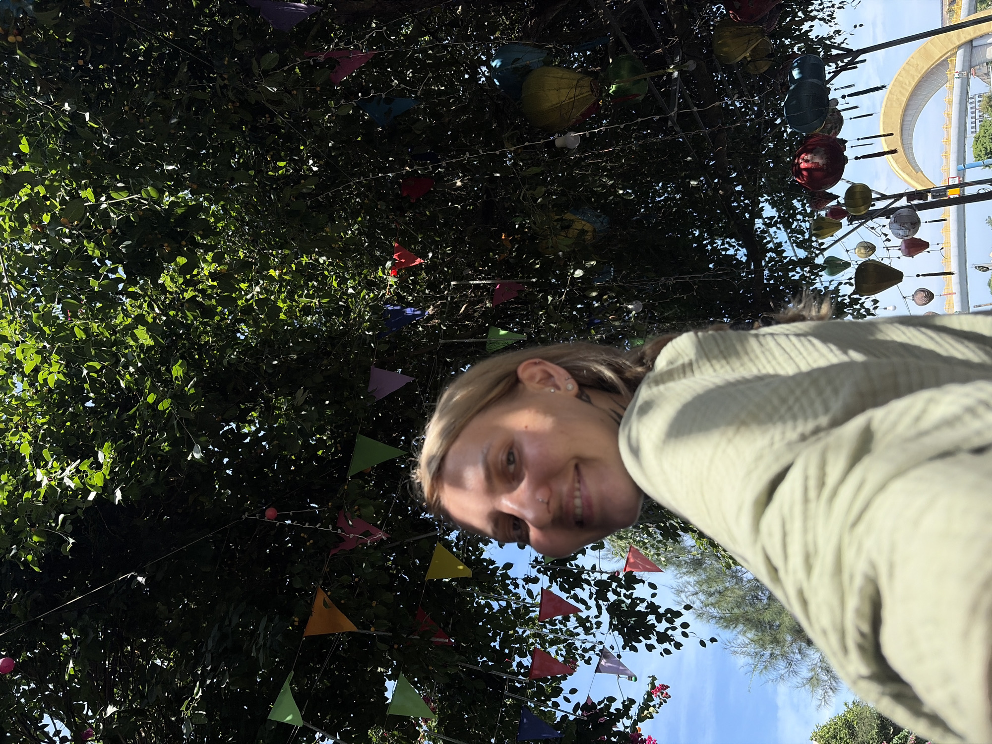 A young woman with blonde hair is featured prominently in the lower right of this bright outdoor selfie, looking up towards the camera with a joyful smile. She is wearing a light-colored, possibly green or beige, ribbed top.
The setting appears to be under a dense canopy of lush green tree leaves, suggesting a warm and sunny climate. Colorful triangular bunting flags, in shades of red, pink, orange, green, blue, and purple, are strung through the foliage, adding a festive atmosphere. Interspersed among the flags and leaves are numerous decorative lanterns of various shapes and colors, including spherical and teardrop designs, some appearing to be light-up decorations.
In the upper right background, against a clear blue sky, a large, distinctive golden archway structure is visible. Below it, a white or light-colored structure, possibly part of a bridge or building, can be seen. The bright sunlight creates dappled light and shadow on the leaves. The time of day is clearly daytime, with bright, clear weather.
The overall impression is one of happiness and celebration in a beautifully decorated outdoor space. The specific lanterns and the architectural archway suggest an East Asian or Southeast Asian location. This aligns with the provided context of Hội An Ward, Vietnam. There is no visible text in the image.