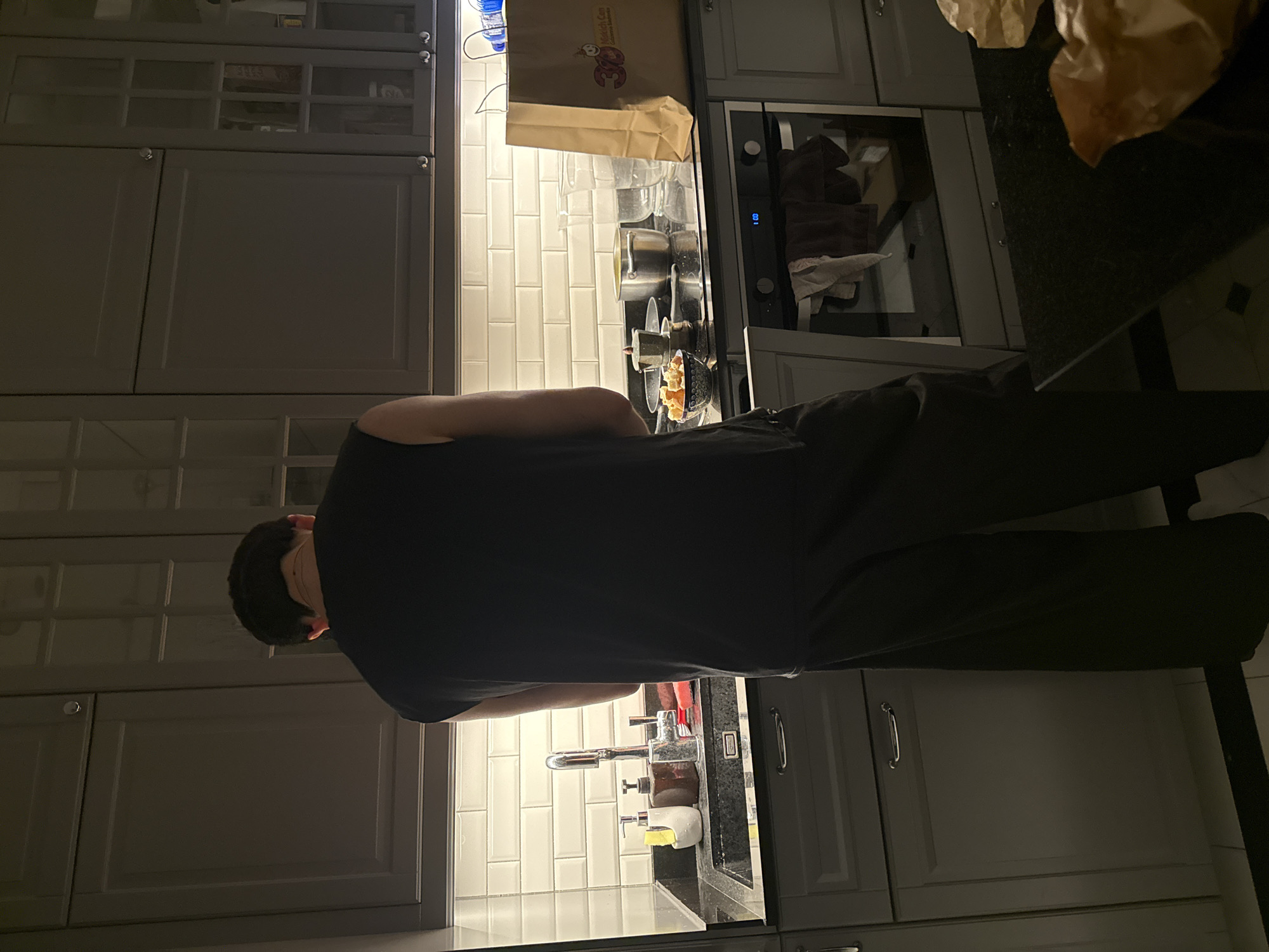 The image depicts a person, viewed from the back, standing in a dimly lit kitchen. The person is wearing a dark, sleeveless shirt and dark pants. They are positioned in front of a kitchen sink, which has a faucet, soap dispensers, and a sponge. To the right of the sink, on the countertop, are several items including pots, bowls containing food, and glasses. A paper shopping bag is also visible on the counter, next to the oven. The kitchen features dark gray cabinets above and below the countertop, with a white tiled backsplash behind the sink and stovetop. The floor is tiled. The overall impression is a domestic scene, possibly at night due to the lighting.