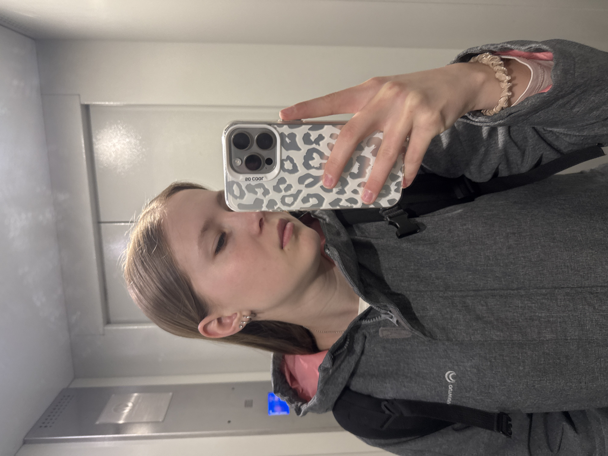 A young person is depicted taking a mirror selfie indoors, in Chaikovsky, Russia. The image is rotated, with the subject's head on the left. They hold a smartphone with a distinctive grey and white leopard print case in their right hand, reflected in the mirror. The subject has light brown hair, wears multiple small silver stud earrings in their left ear, and a thin silver necklace. They are dressed in a grey jacket with a pink inner lining, and a black backpack strap is visible over their right shoulder. A light-colored, textured hair tie or bracelet is worn on the right wrist. The person's expression is neutral, with eyes slightly downcast.
The setting appears to be an elevator or a similar enclosed space with light-colored, plain walls. A metal control panel with a glowing blue light is visible in the reflection on the lower left wall, reinforcing the elevator context.
Visible text includes "SO COOR" (reversed) on the phone case. On the grey jacket, a partial logo appears as "OALMBIA" (reversed), suggesting the brand "COLUMBIA". There are no specific cues for time of day or weather as the scene is indoors.