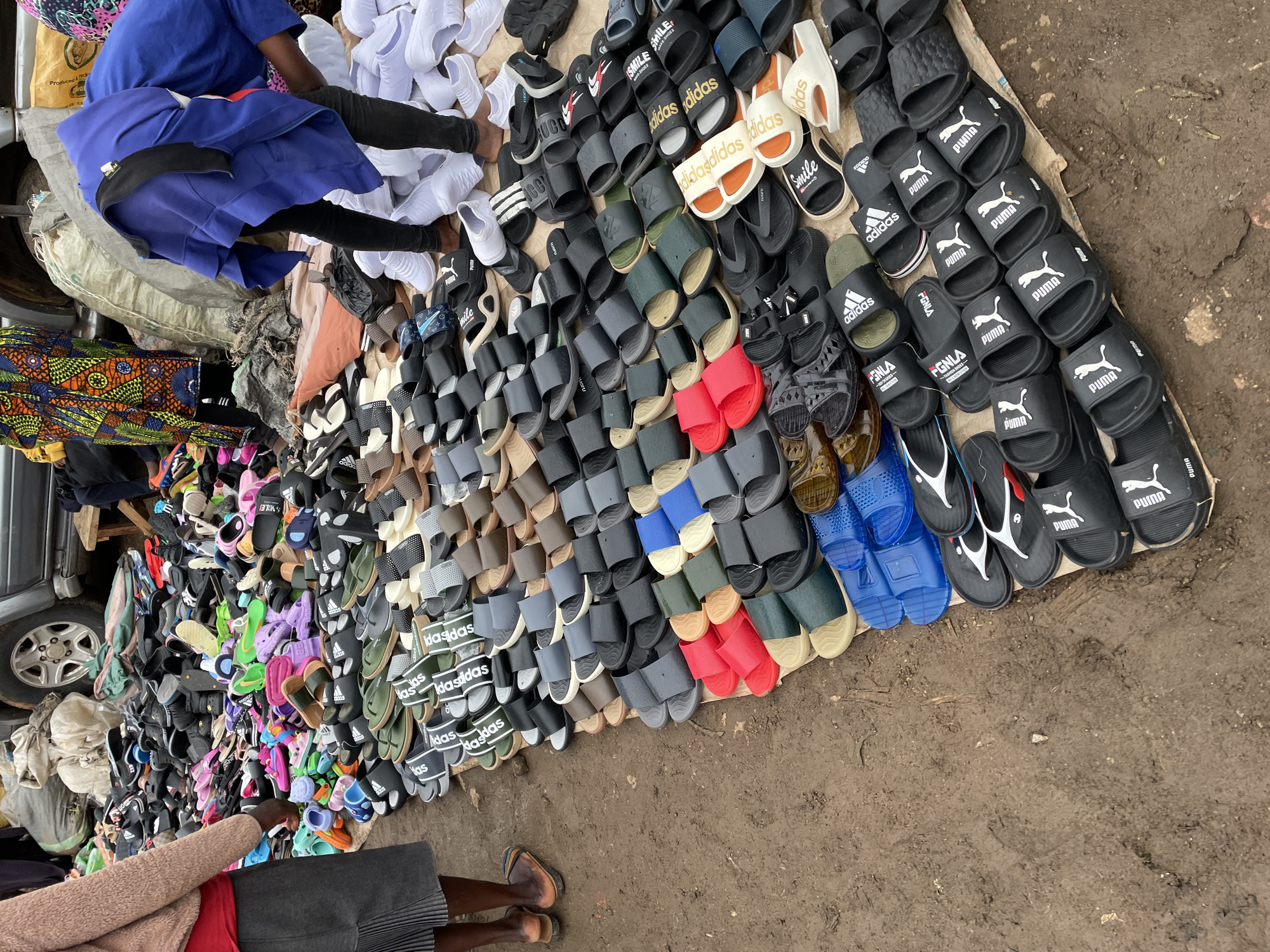 Captured in the Nigerian city of Jos, an outdoor market scene unfolds on a neutral-toned, possibly concrete, surface. A dense collection of sandals and slides dominates the frame, organized in rows. Various brands are visible, including "adidas," "PUMA," and "Smile," in a variety of colors and styles.
Two individuals appear in the scene. One individual, wearing a blue top and black bottoms, is positioned near the top of the frame and seems to be sorting or organizing the sandals. Another individual, clad in a red top, gray bottoms, and a tan sweater, is seen near the bottom of the frame, perhaps browsing the selection.
A car is parked adjacent to the market stall on the left, and a piece of colorful patterned textile rests nearby, possibly part of the vendor's goods. The overall environment suggests a busy marketplace in Jos.