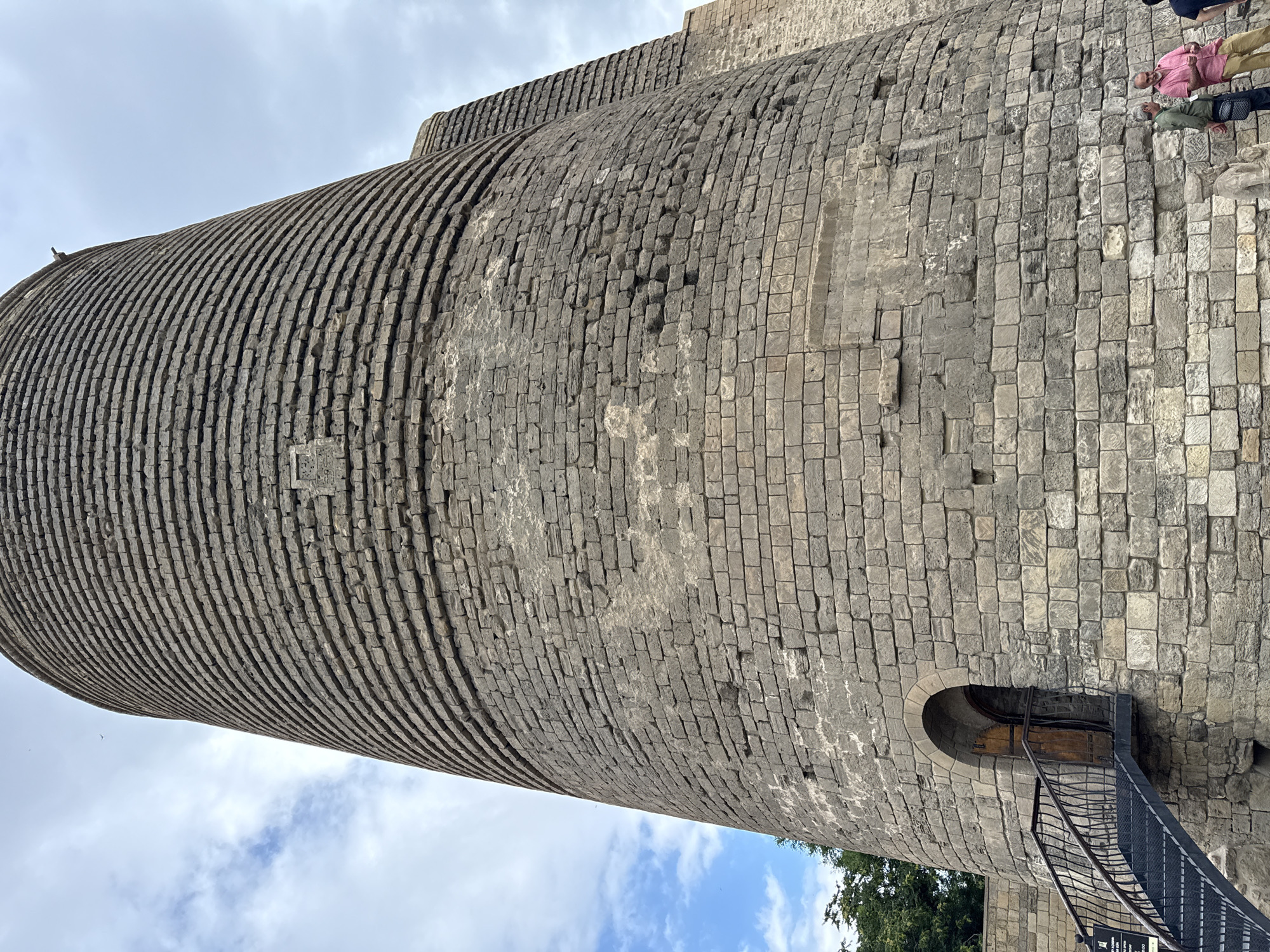 The image shows the Maiden Tower, a UNESCO World Heritage site in the Old City of Baku, Sabail Raion, Azerbaijan. The tower is a cylindrical stone structure with a distinctive conical top, though the top is not fully visible in this shot. The stone blocks are aged and weathered, with some areas showing signs of wear and repair. A metal staircase with a black railing leads to an arched wooden doorway on the lower left side of the tower. In the distance, another similar tower structure is partially visible to the right, indicating a complex of towers. Two men are standing near the base of the main tower on the right, looking at something out of frame. One man is wearing a pink shirt and beige pants, and the other is wearing a green shirt and dark pants with a bag slung over his shoulder. The sky above is partly cloudy, with patches of blue sky visible. Green trees are seen on the far left, behind a stone wall. The overall scene depicts an ancient architectural landmark under a partly cloudy sky.