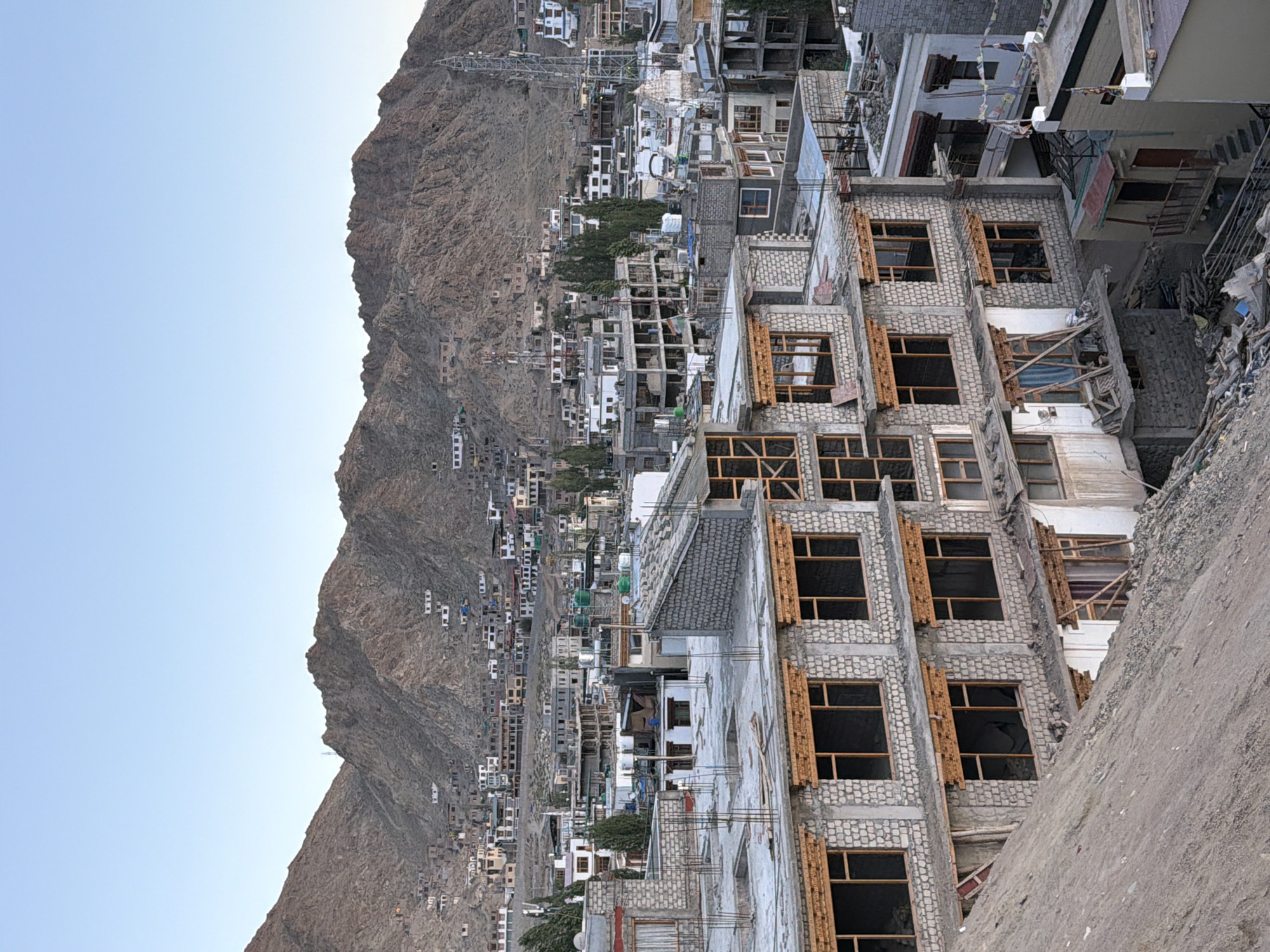 The image depicts a hillside view of the city of Leh, India, with a focus on buildings under construction and the surrounding mountainous landscape. In the foreground, a partially built concrete structure with exposed rebar and unfinished stone facades dominates the scene. The building has multiple floors with empty window frames and wooden detailing above some of the windows. To the left, a steep, dusty slope rises from the base of the constructed building.
In the mid-ground, the city of Leh unfolds, characterized by a dense cluster of buildings. Many of these structures appear to be multi-story, with a mix of stone and concrete construction. Some buildings are complete, while others show signs of ongoing construction or renovation, with visible scaffolding and unfinished sections. Several prominent features stand out, including tall communication towers, green water tanks on rooftops, and a small white temple or stupa with a golden finial. The buildings extend up the gentle slope of the mountains that form the backdrop.
The background is dominated by arid, rocky mountains with sparse vegetation. The mountains are terraced in some areas, suggesting human activity or natural formations. The sky is clear and light blue, indicating daytime, likely late afternoon or early evening due to the soft lighting. There are no people or animals visible in the frame. The overall impression is one of urban development in a rugged, mountainous environment.