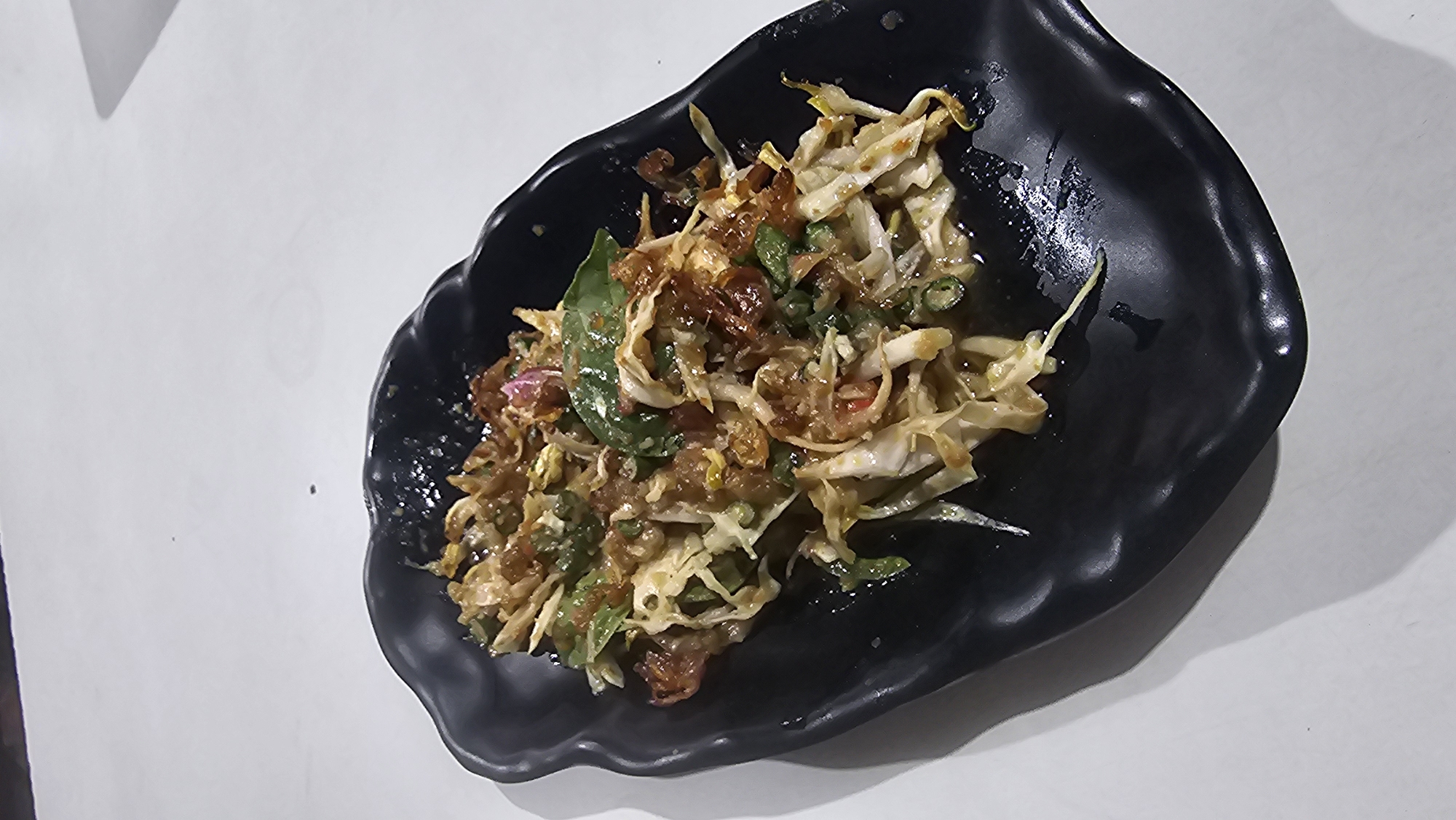 A close-up, high-angle shot shows a dark, wavy-edged plate filled with a vibrant salad. The salad is a mix of shredded cabbage, green onions, what appears to be fried shallots, and herbs, all tossed in a glistening dressing. The plate sits on a white, flat surface, casting a shadow to the left. The environment suggests an indoor setting, possibly a restaurant in Pasirhalang, Indonesia, given the context. There are no people visible in the image.