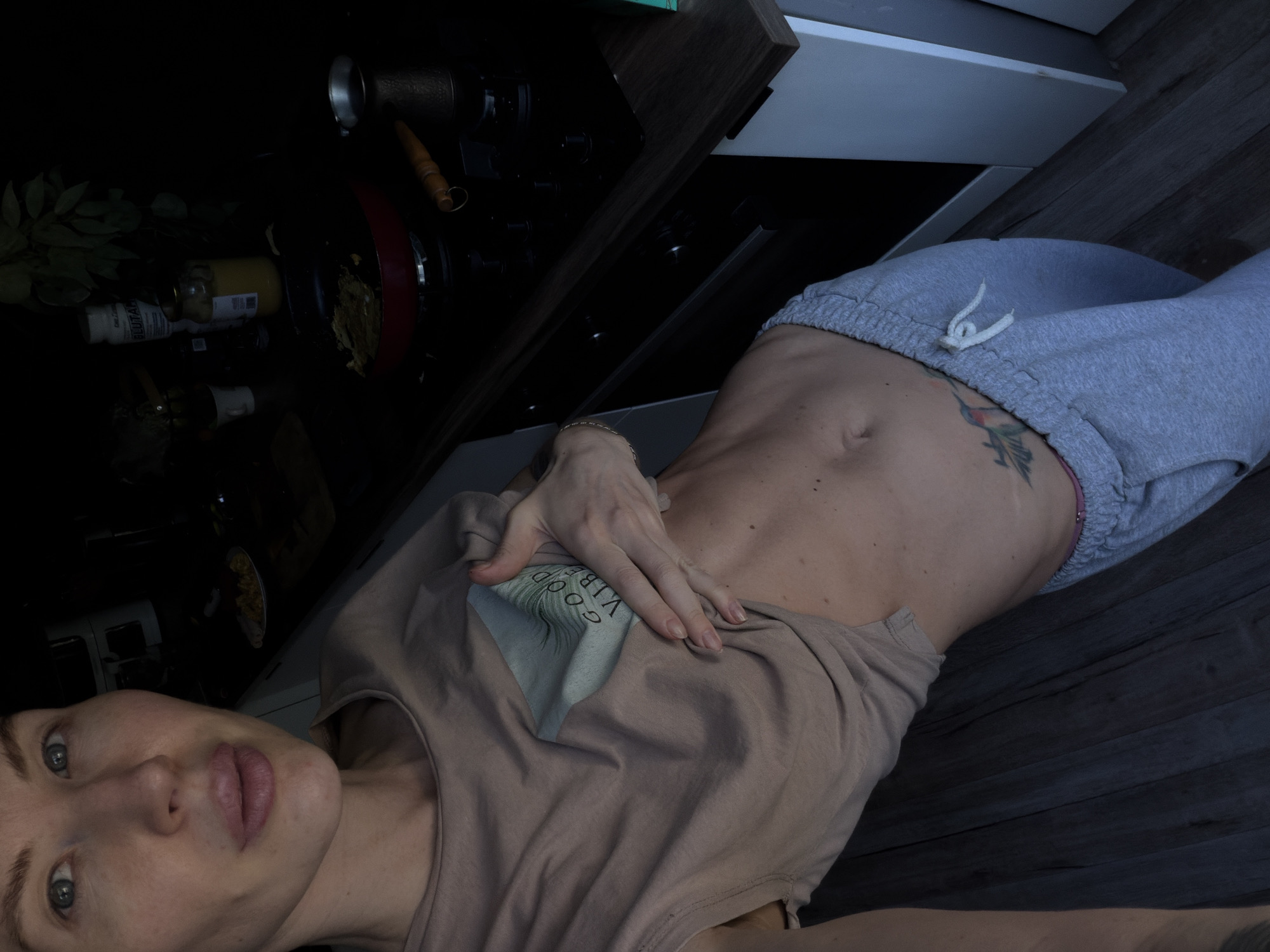 A fair-skinned person is lying on a wooden floor in a domestic kitchen setting. The person is wearing grey sweatpants and a beige t-shirt with the phrase "GOOD VIBES" printed on it. Their right hand is resting on their chest. The kitchen background features a stove, an open cabinet, and various kitchen items, including bottles and a pan with food. The image is taken from a low angle. There is no information to determine the location is Perm, Russia.