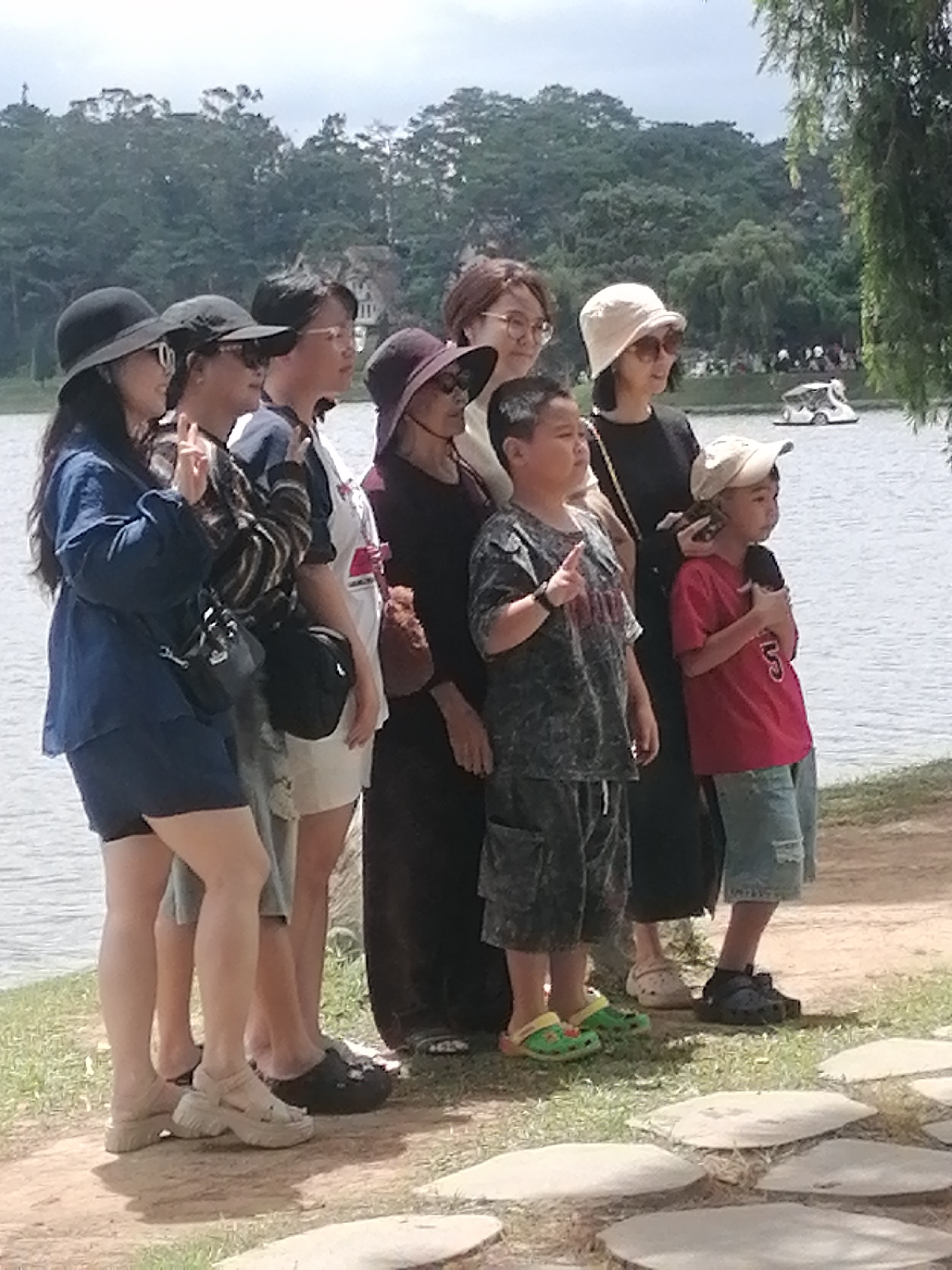 A group of six people, including two young boys and four women, stand by the edge of a lake on a sunny day. They are all looking in the same direction, towards the water. The group is composed of individuals of Asian descent.
The people are dressed casually. The women are wearing various hats and sunglasses. The children are also in casual attire. One of the boys is wearing a dark, patterned t-shirt and shorts, while the other is in a red t-shirt and denim shorts.
The environment appears to be a park or recreational area. In the background, there is a body of water, a treeline, and some distant buildings. A small boat is visible on the lake. The ground in the foreground is a mix of grass and a paved pathway made of large, flat stones.
This scene is likely located in the city of Da Lat, Vietnam, a popular tourist destination known for its lakes, pine forests, and French colonial architecture. The presence of a lake, lush greenery, and a relaxed atmosphere are characteristic of many attractions in Da Lat, such as Xuan Huong Lake or the lake at the Valley of Love.
