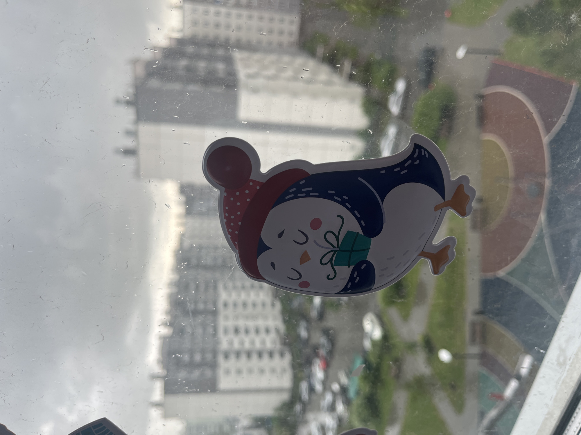 The image presents a close-up, rotated view of a window pane with a decorative sticker, looking out onto a blurry urban or suburban environment. The window is covered in numerous small raindrops and smudges, indicating it's either raining or has recently rained.
The primary subject is a cartoon penguin sticker, placed on the window. The penguin is depicted lying on its back, oriented with its head towards the top-left of the frame. It wears a red knitted hat adorned with white polka dots and has its eyes closed, suggesting a happy or peaceful expression. The penguin holds a small green gift box tied with a lighter green bow. Its body features dark blue plumage on top, a white belly, and orange beak and feet.
Beyond the rain-streaked glass, the background is significantly blurred, creating a shallow depth of field with the sticker in sharp focus. Tall, light-colored buildings, possibly residential or commercial, are visible through the haze and raindrops. Below these structures, patches of green vegetation and what appears to be a street or parking lot with indistinct vehicles can be discerned. The scene suggests a daytime view from an elevated position within a building, looking out over a city or town during inclement weather. No visible text is present in the image.