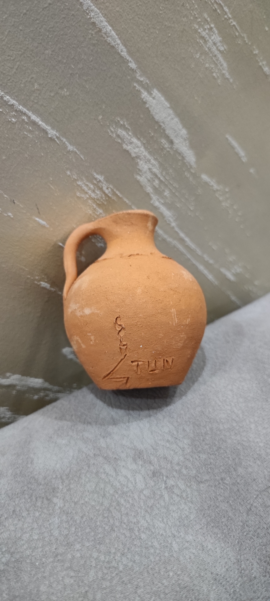 A small, tan, clay pot sits on a gray, textured surface. The pot has a handle and is inscribed with the letters "TUN" and an abstract design. The background consists of a textured wall. The location of Yerevan, Armenia is assumed from the inscription "TUN," but is not visibly confirmed.