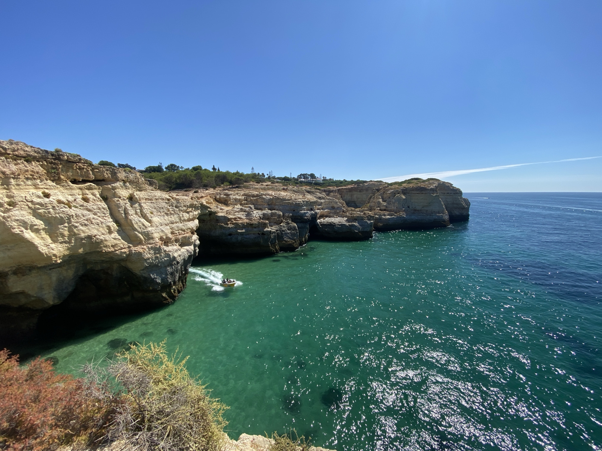A bright, sunny day is depicted with a clear blue sky above a rocky coastline. The foreground features rugged, light-colored cliffs with visible erosion and some sparse vegetation. The sea is a vibrant turquoise, so clear that the seabed is visible in shallower areas. A small yellow boat with a few people aboard is speeding across the water, leaving a white wake. In the distance, the coastline continues with similar rock formations and some greenery on the clifftops, suggesting a natural, undeveloped area. The overall scene captures the beauty of the Portuguese coast in Lagoa e Carvoeiro.