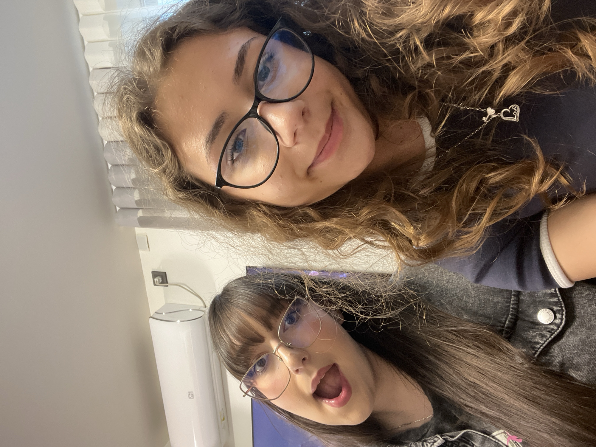 Two young women are posing indoors for a selfie. The woman in the top portion of the frame has long, curly, light brown hair, and wears black-framed glasses. She is smiling slightly and has a heart-shaped necklace around her neck. The woman in the lower portion of the frame has straight, dark brown hair with bangs and wears gold-framed glasses. Her mouth is open in a surprised or excited expression. The background includes a white wall, a window with blinds, an air conditioner unit, and an electrical outlet. The location is not indicated within the image.