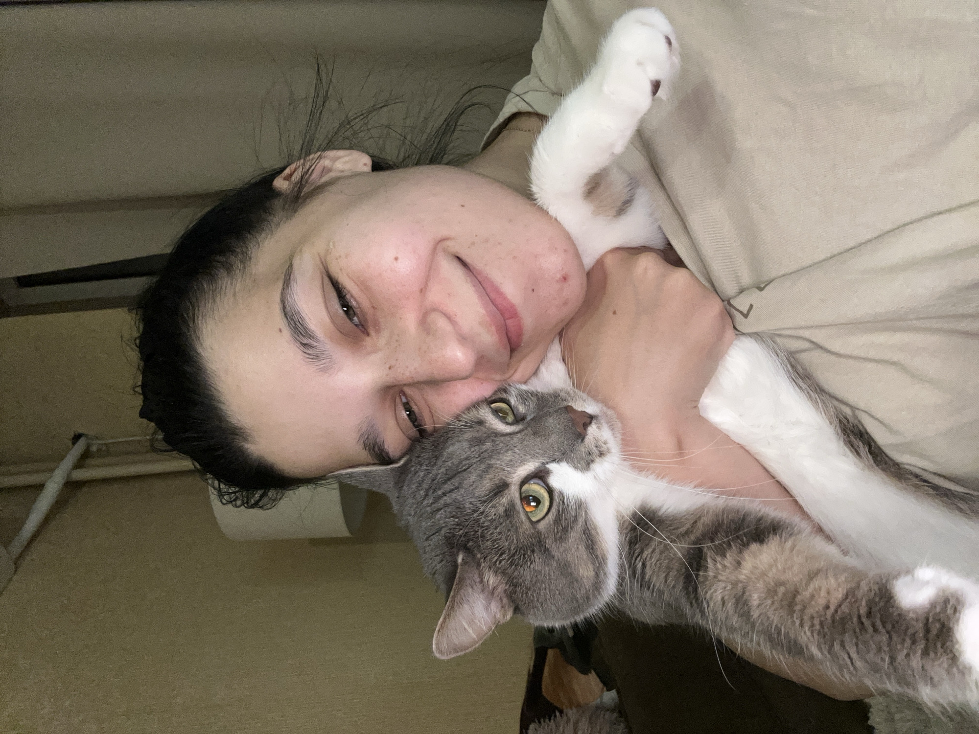 An indoor, eye-level shot shows a person cuddling a cat. The image is oriented with the subjects lying horizontally, with the person on the left and the cat on the right, both looking generally towards the camera, though the cat's gaze is directed slightly upwards and to the right.
The person, possibly female, has dark hair pulled back and a fair complexion with visible freckles on her face. She is smiling gently, showing a sense of contentment and affection. Her left hand is visible, holding the cat close to her body. She is wearing a light-colored, possibly beige or cream, top.
The cat is a domestic short-haired breed, predominantly grey and white, with distinct green-amber eyes. Patches of tan or brown fur are also visible, particularly on its body. Its white paws are prominent, with one near the person's face. The cat appears relaxed and comfortable, resting against the person.
The background is simple and somewhat indistinct, featuring light beige or tan walls, suggesting a home or apartment setting in Sofia, Bulgaria. In some cropped views, faint elements like a pipe or cable and a roll of paper are visible in the upper background. The lighting is soft and ambient, characteristic of an indoor environment without direct harsh light. The overall mood of the image is one of warmth, affection, and companionship between the person and their pet. No visible text is present in the image.
