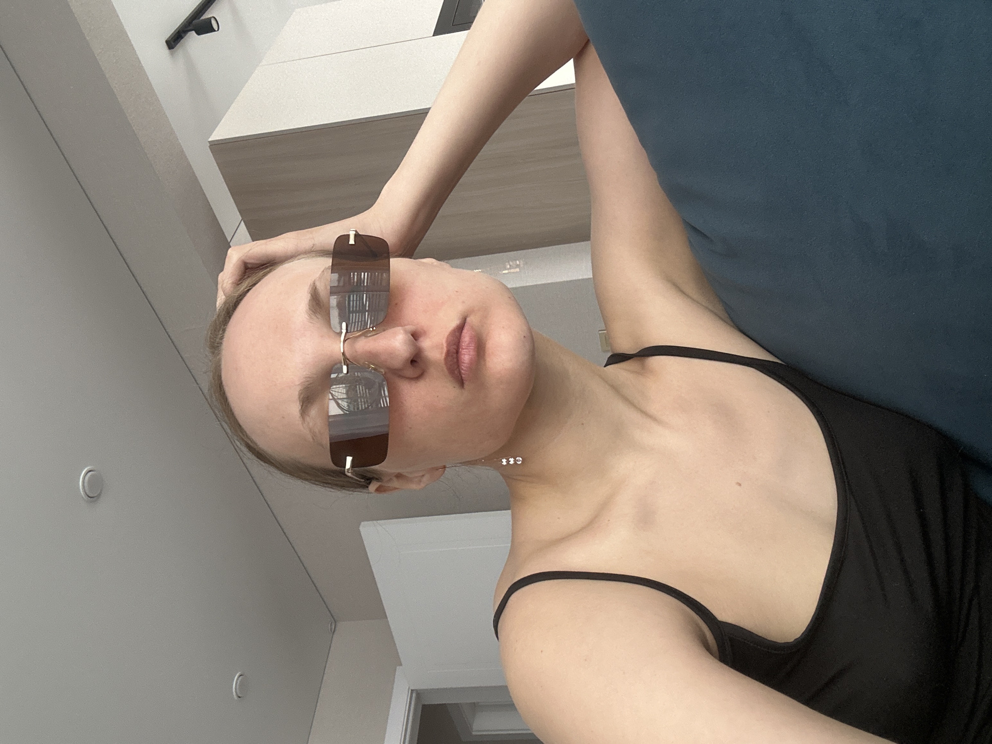 A woman with fair skin and light brown hair is indoors, wearing sunglasses and a black tank top. Her left hand is on her head. She is leaning against a blue cushion or sofa. The background shows a white wall, a white cabinet, a dark-colored light fixture, and a white door frame. The location is unknown.