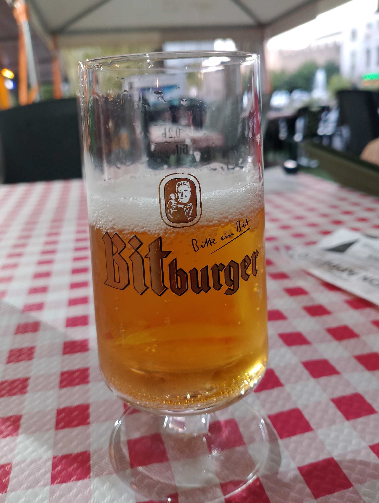 A close-up, eye-level, vertical shot shows a glass of Bitburger beer. The glass is mostly full, with a frothy head of foam on top, and the beer itself is a clear golden-yellow color. The glass has a distinct logo with a man's profile and the words "Bitburger" and "Bitte ein Bit" printed in brown lettering. The glass is resting on a red and white checkered tablecloth, which covers a table outdoors, likely at a restaurant or beer garden. In the blurred background, tents, other tables, and some greenery are visible, suggesting an outdoor dining setting. The image was taken in the city of Rieti, Italy.