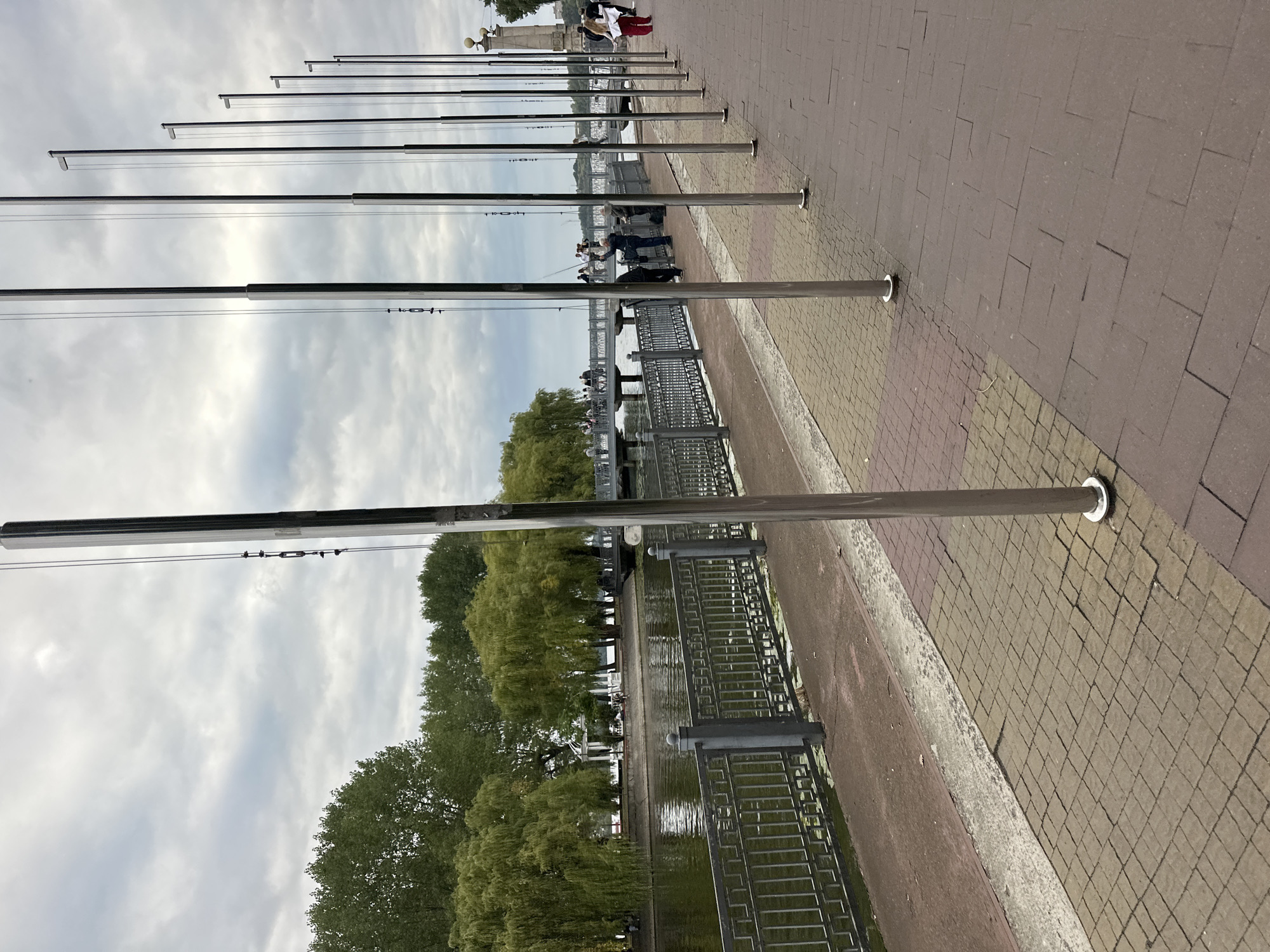 This is an eye-level, medium shot of a waterfront promenade in Ternopil, Ukraine, under a cloudy sky. In the foreground, a row of tall, slender, metallic flagpoles stands to the right, reflecting the overcast sky. The promenade itself is paved with reddish-brown and beige square bricks. To the left of the flagpoles, a metal railing with a decorative pattern separates the promenade from a body of water, likely Lake Ternopil. Beyond the railing, a stone embankment lines the water, with lush green trees, including weeping willows, bordering the far side. A bridge with people on it spans the water in the mid-ground, connecting to a white gazebo-like structure visible in the distance. Several individuals are scattered along the promenade and the bridge, with one person appearing to fish from the bridge. Another person, dressed in red, walks away from the camera on the right side of the promenade. The overall scene depicts a peaceful, public outdoor space in a city, characterized by its natural elements and urban infrastructure.