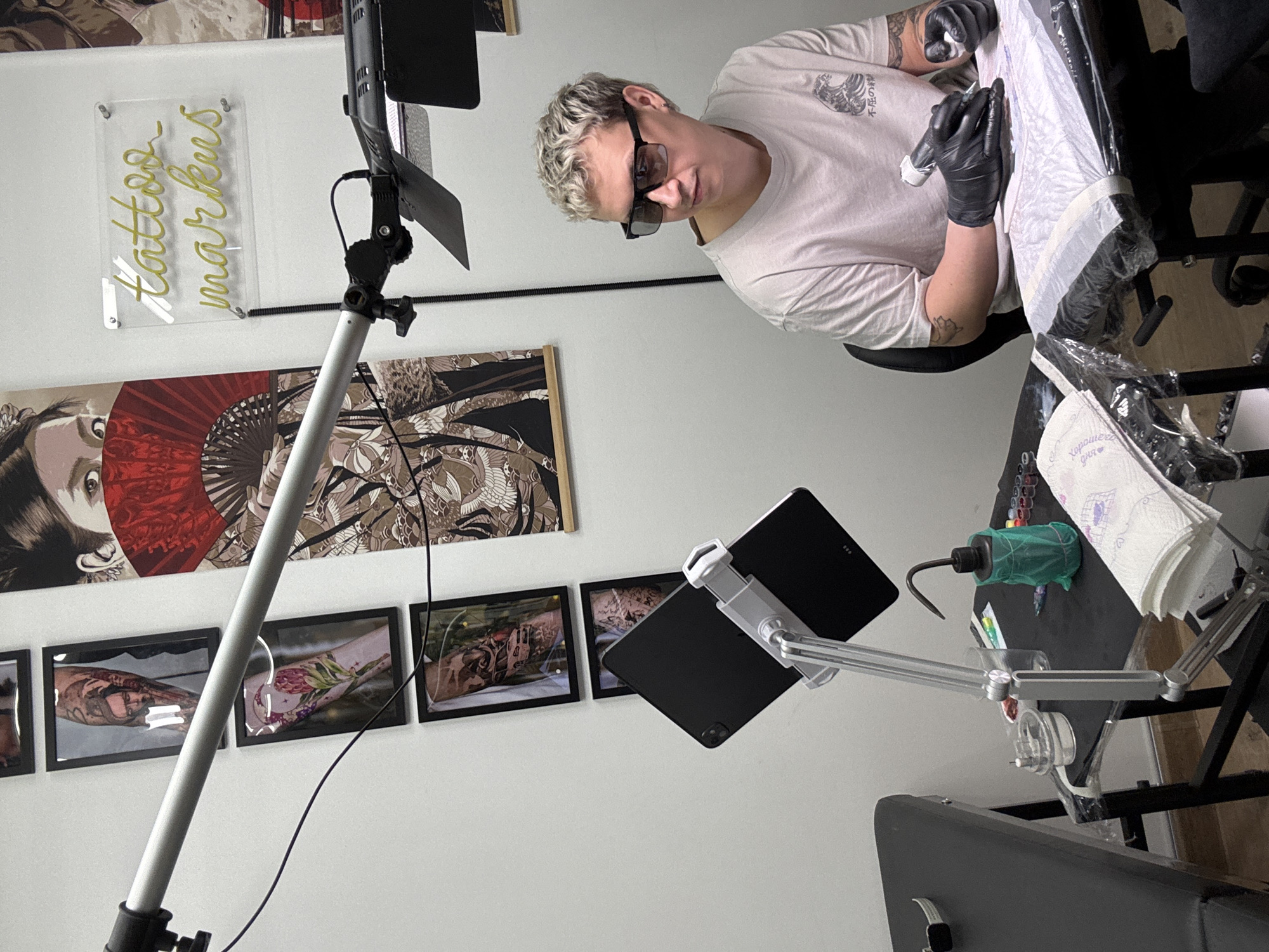 This image captures a tattoo artist at work in a professional tattoo studio in Chaikovsky, Russia. The artist, a person with short, light-colored curly hair and glasses, is depicted from a slightly elevated, side-angle perspective, intently focused on tattooing a client's arm. The artist wears a light-colored t-shirt with a subtle wave graphic on the chest and black protective gloves, holding a tattoo machine over the client's plastic-wrapped arm. Tattoos are visible on the artist's own arms. The artist's expression is concentrated and professional.
The studio environment is well-equipped and organized. An adjustable overhead light fixture with barn doors provides illumination to the workspace. To the artist's right, a tablet, likely an iPad, is mounted on a flexible arm. The work surface directly in front of the artist is covered in protective plastic and holds various tattooing essentials, including a green squeeze bottle, several small bottles of ink, and a roll of paper towels. A small piece of paper on the table displays the Russian text "Хорошего дня ❤️" (Have a good day ❤️).
The background wall is adorned with several decorative elements. A large print featuring a woman with a red fan is prominent, and below it, a series of smaller framed pictures showcase various tattoo designs and tattooed arms. Above these decorations, a clear acrylic sign with yellow neon-style cursive text reads "tattoo markus", likely indicating the studio's name or the artist's alias. The scene suggests a clean, organized, and dedicated workspace, brightly lit indoors.