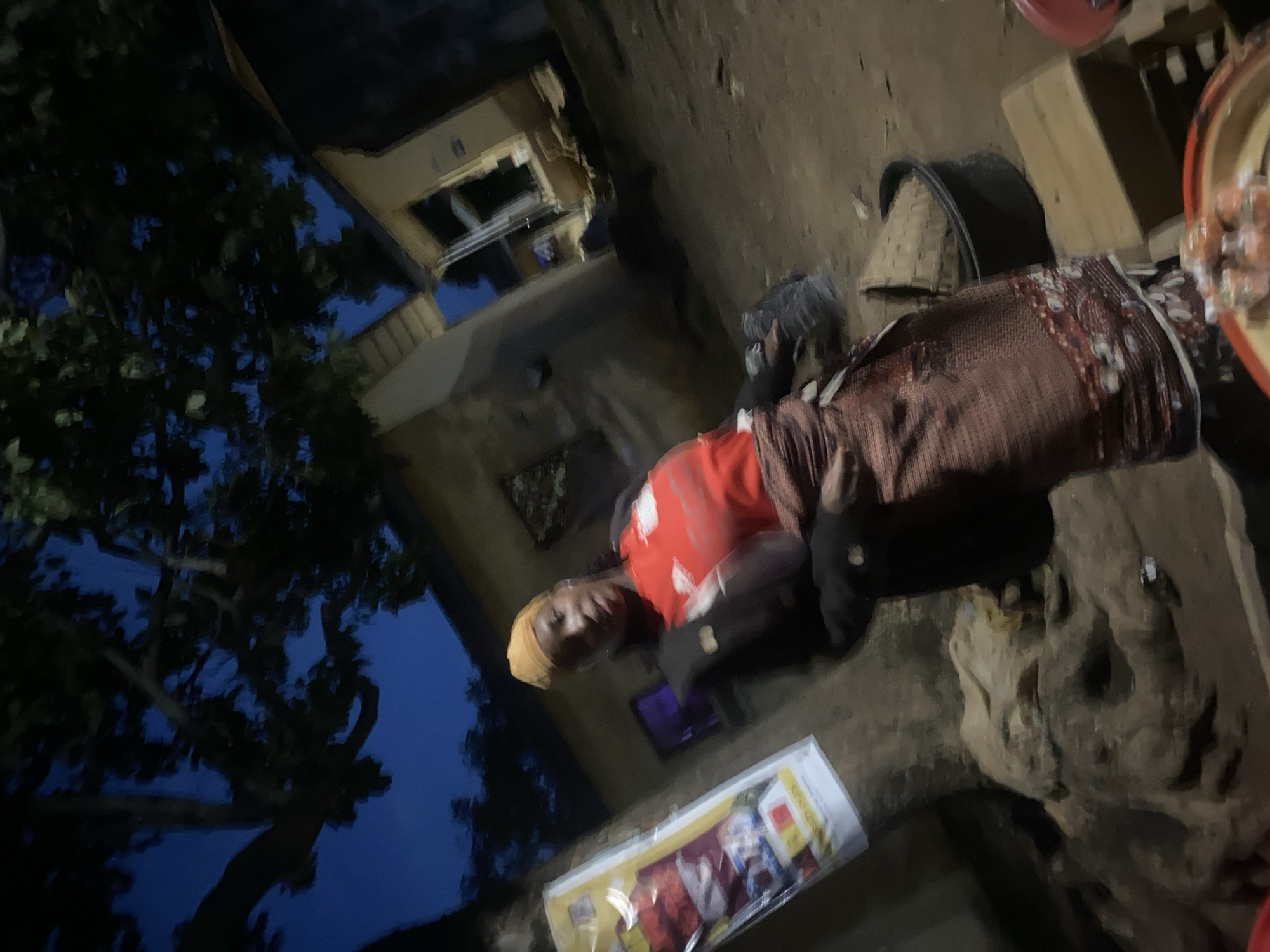 A woman wearing a yellow headscarf and a red shirt is standing outdoors at dusk in Rafin Par, Nigeria. She has a brown wrap around her waist and is holding a black bag. In the background, there are buildings and trees, and the sky is dark blue, indicating it is nighttime. The ground is dirt and there are various objects around, including a large black tub, a wooden crate, and a pile of sacks. A poster is attached to a tree on the left side of the image. The scene appears to be a marketplace or a street in a residential area.