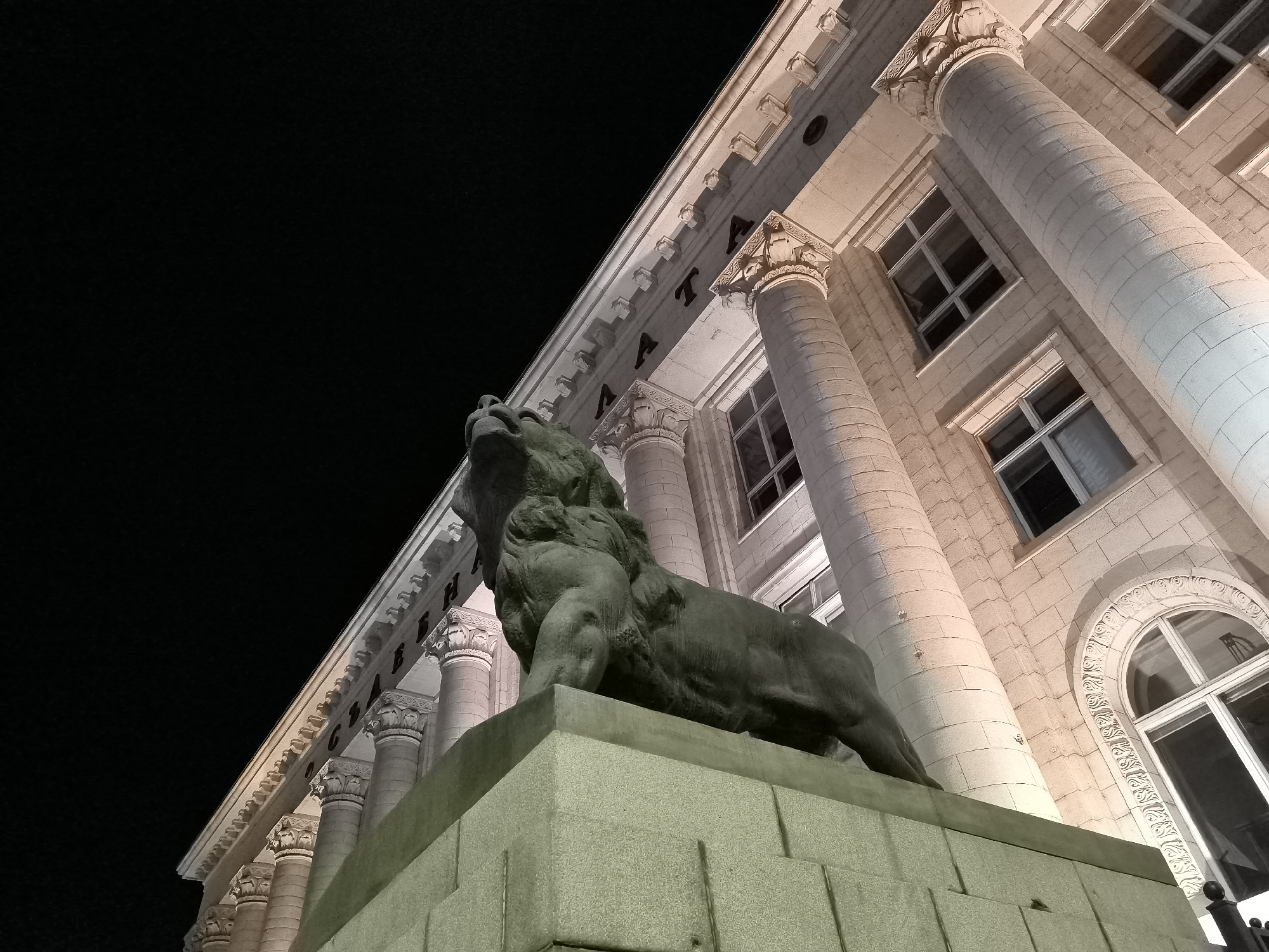 A low-angle, close-up shot captures a monumental stone lion sculpture positioned on a tiered pedestal, facing left with its head tilted upwards and mouth open in a roar. The sculpture is situated in front of a grand, classical-style building illuminated at night. The building features a facade of light-colored stone blocks, adorned with a series of large, fluted Corinthian columns. Above the columns, a decorative cornice runs along the building's upper edge, punctuated by ornate carvings and a series of small, rectangular protrusions. Several large, multi-paned windows are visible between the columns, some appearing dark, others reflecting the building's illumination. The dark night sky forms the backdrop to the scene. The sculpture and building are bathed in a warm, artificial light, highlighting the textures of the stone and casting shadows. The overall impression is one of solemnity and grandeur. This is the Lion Monument in front of the National Palace of Culture in Sofia, Bulgaria.