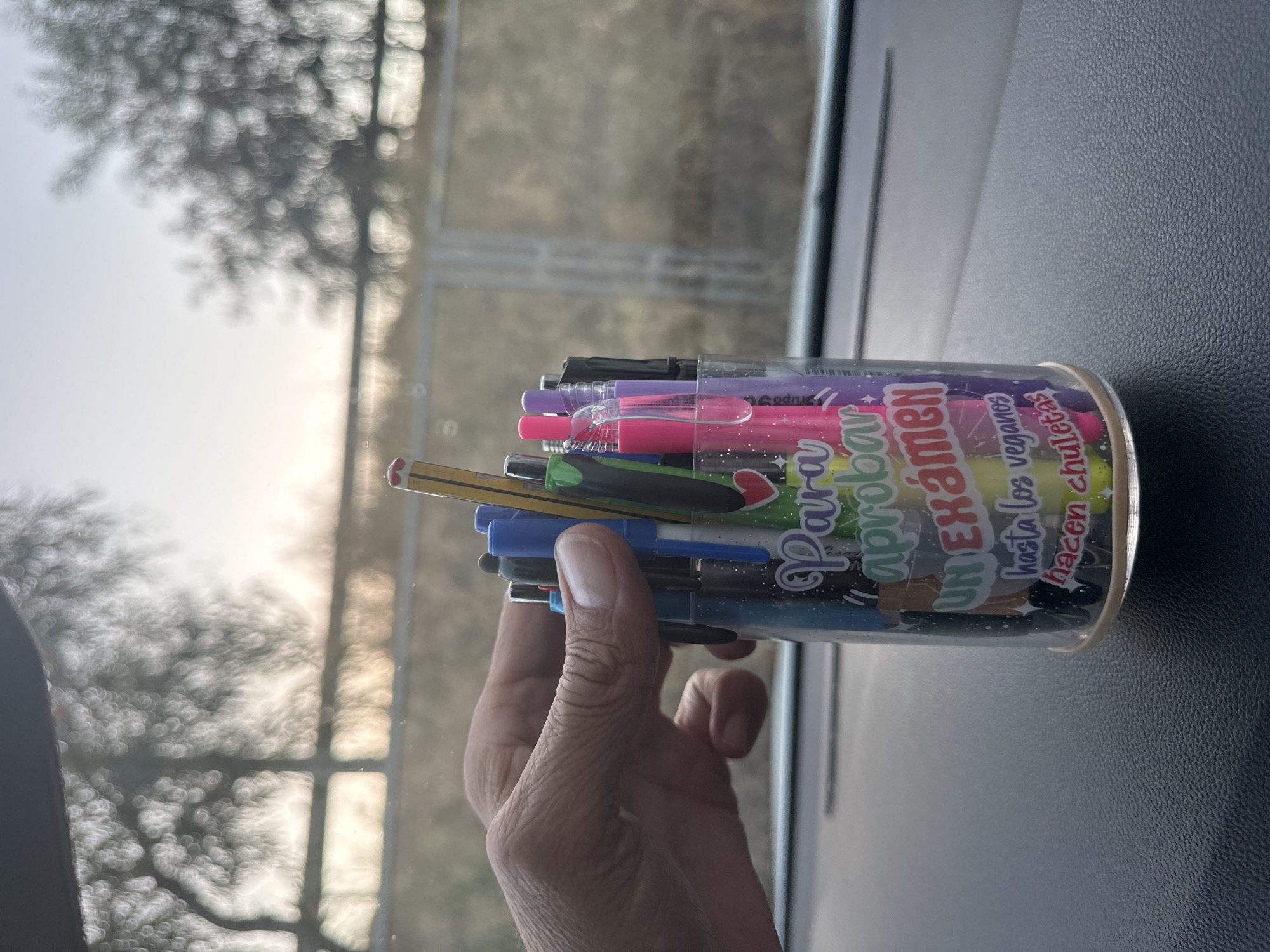 A hand is reaching into a clear plastic pen holder filled with colorful pens and a pencil. The holder has stickers with Spanish text that reads "Para aprobar un examen hasta los veganos hacen chuletas" which translates to "To pass an exam, even vegans make cheat sheets." The pen holder is placed on a dark, textured surface, likely the dashboard of a vehicle, with a blurred background of trees and sky, suggesting an outdoor setting, possibly on the outskirts of the city of Murcia, Spain. The lighting indicates it is daytime, possibly late afternoon or early morning.