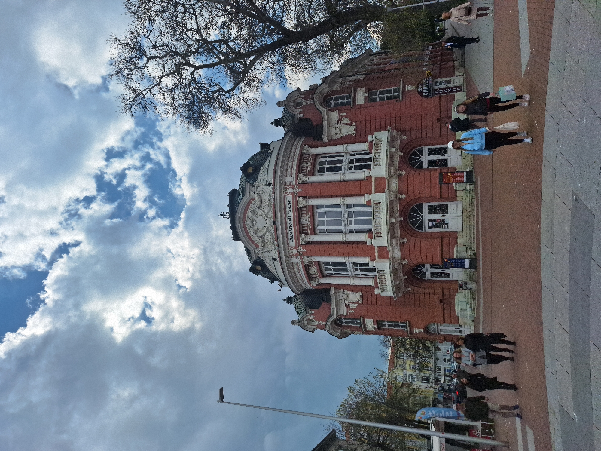 The image captures a view of the Dramatic Theatre "Stoyan Bachvarov" in Varna, Bulgaria. The architectural style is distinct, featuring a red brick facade with white ornamental details and a rounded section. People are visible on the pavement in front of the theatre. The sky is partly cloudy, with a tall, leafless tree visible. A street light post is present on the left.