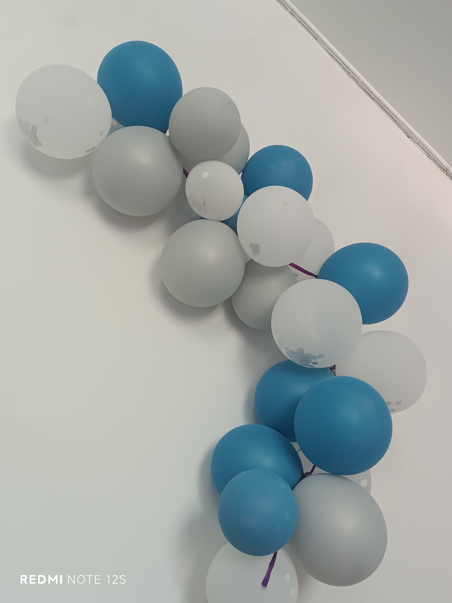 A close-up, vertical, eye-level shot captures a string of balloons in varying shades of blue, white, and gray, arranged in a curving line against a plain white wall. The balloons are primarily smooth and round, though some of the white ones have subtle, light gray floral patterns. A thin, purple ribbon is visible, connecting some of the balloons. The upper right corner of the image shows a slight shadow cast by the balloons and a sliver of a ceiling line, indicating an indoor setting. The bottom left corner of the image features the text "REDMI NOTE 12S" in white. The scene is devoid of any people or animals. The specific location of Kara-Bulak, Kyrgyzstan, is not discernable from the visual elements within the image.