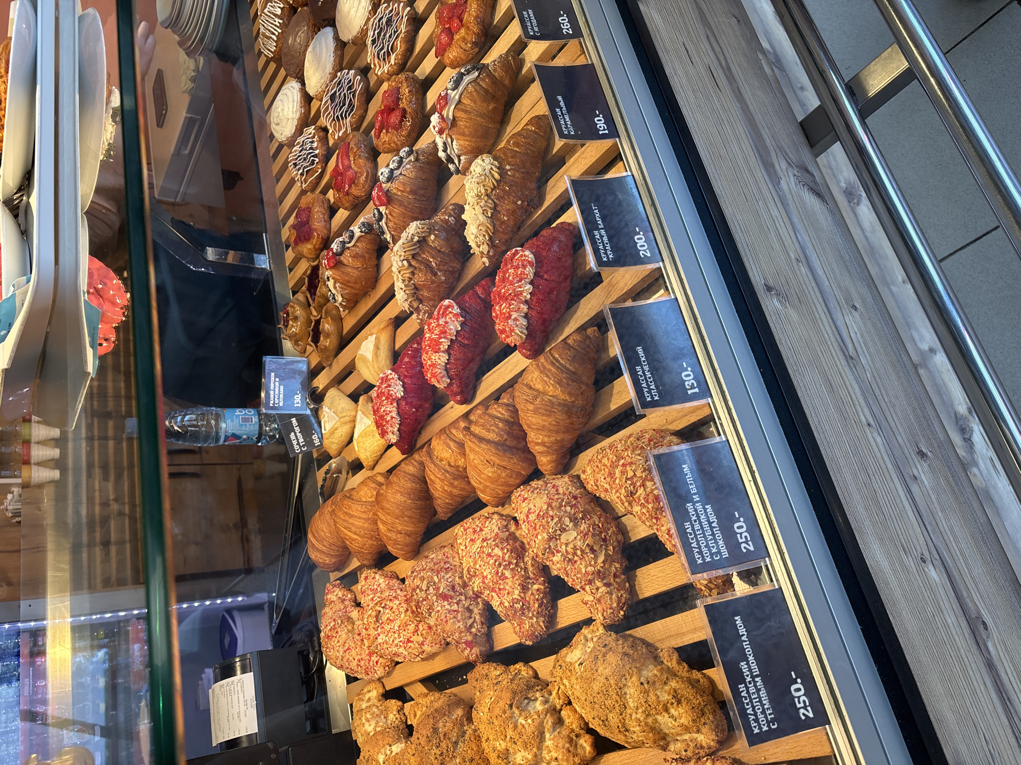 This image captures a vibrant display of pastries in a bakery or cafe in Rusanovo, Russia. The scene features a glass-fronted display case with multiple wooden shelves, showcasing a wide assortment of freshly baked croissants and other sweet treats. Many croissants are decorated with various toppings, including dark and white chocolate drizzles, fresh berries like strawberries and blueberries, sliced almonds, and colorful crumbs resembling red velvet or other sweet coatings. Some pastries appear plain or with a simple glaze.
Small black price tags with white Cyrillic text and numerical prices are visible in front of the items. These tags identify several types of croissants: "КРУАССАН С ЯГОДАМИ" (Croissant with berries) for 260 rubles, "КРУАССАН КАРАМЕЛЬНЫЙ" (Caramel croissant) for 190 rubles, "КРУАССАН "КРАСНЫЙ БАРХАТ"" (Croissant "Red Velvet") for 200 rubles, "КРУАССАН КЛАССИЧЕСКИЙ" (Classic croissant) for 130 rubles, "КРУАССАН КОРОЛЕВСКИЙ С КЛУБНИКОЙ И БЕЛЫМ ШОКОЛАДОМ" (Royal croissant with strawberries and white chocolate) for 250 rubles, and "КРУАССАН КОРОЛЕВСКИЙ С ТЕМНЫМ ШОКОЛАДОМ" (Royal croissant with dark chocolate) for 250 rubles. Other visible items include a "РЖАНОЙ ПИРОЖОК С БРУСНИКОЙ И ЯБЛОКАМИ" (Rye pie with lingonberries and apples) for 130 rubles and "СОЧЕНЬ С ТВОРОГОМ" (Socheń with cottage cheese) for 160 rubles. A bottle of water is also visible on an upper shelf, priced at 160 rubles. Stacked white plates and serving tongs are seen to the left, ready for customers. Reflections on the glass hint at the surrounding environment but no people are clearly visible. The overall impression is one of an inviting and well-stocked pastry counter.