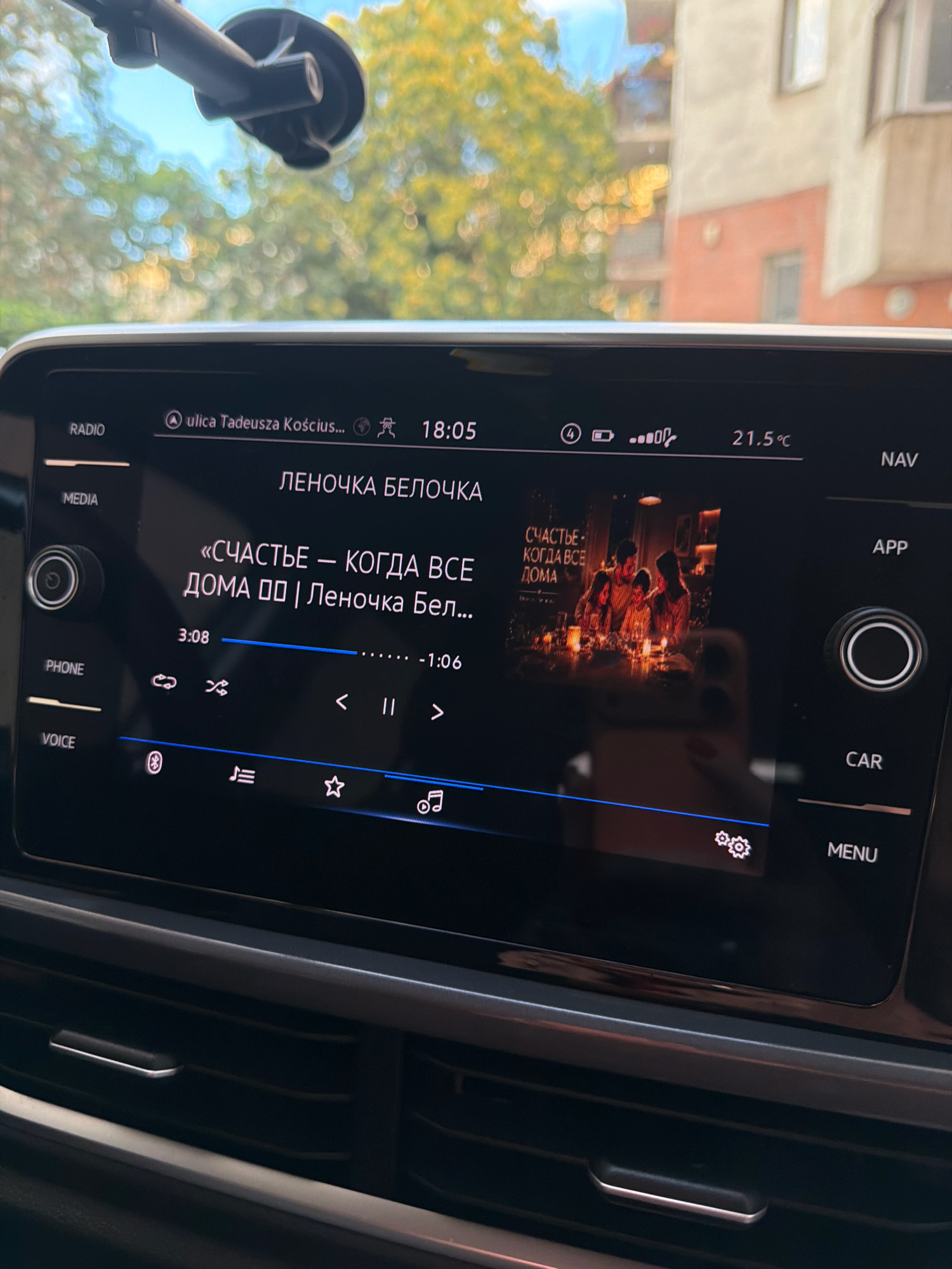 The image shows a car's infotainment system displaying a song titled "СЧАСТЬЕ — КОГДА ВСЕ ДОМА" (Happiness - When Everyone is Home) by "Леночка Белочка" (Lenochka Belochka). The time displayed is 18:05, and the temperature is 21.5°C. The navigation system indicates a location at "ulica Tadeusza Kościuszki," which is a street in Wrocław, Poland. The screen also shows various icons related to music playback, phone connectivity, and settings. The background of the image, visible through the car's windshield, shows trees with yellowing leaves and a building, suggesting an outdoor setting.