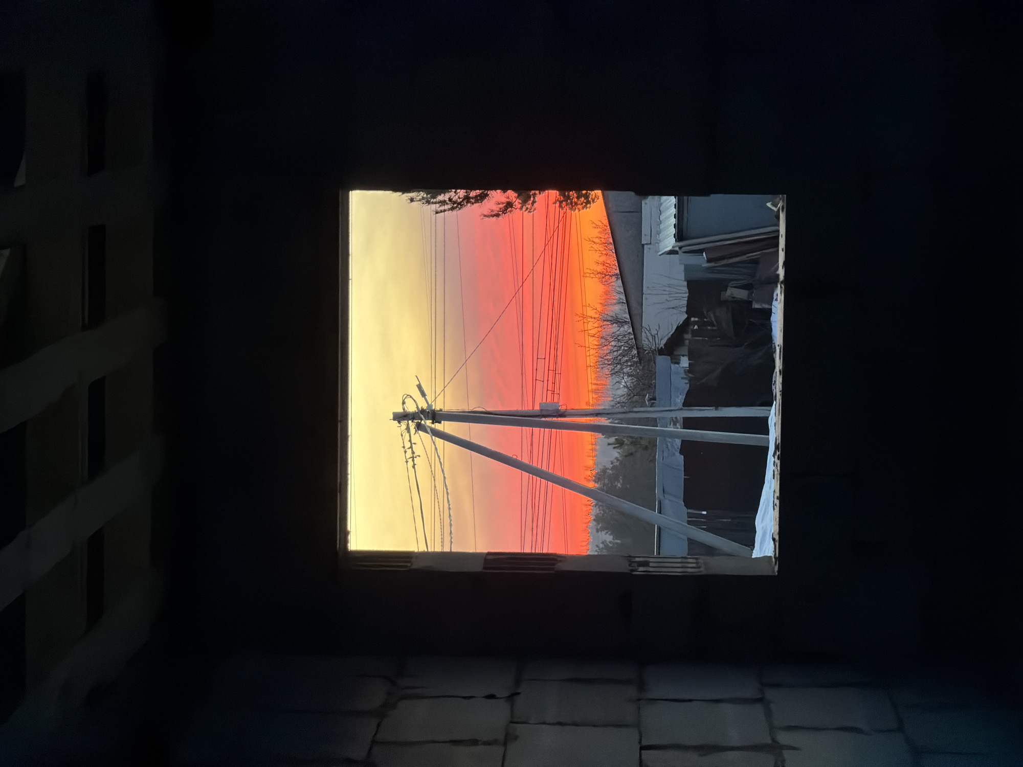 A low-angle, medium shot depicts an intense sunset visible through a square window opening of an unfinished brick building. The interior walls of the building are dark, creating a stark contrast with the vibrant sky. Outside the window, power lines and poles are visible, silhouetted against the gradient of colors in the sky, transitioning from yellow at the horizon to orange and red higher up. To the right, parts of neighboring structures can be seen. The location is most likely Chaikovsky, Russia, as implied in the prompt. The bricks on the floor show the interior is under construction.