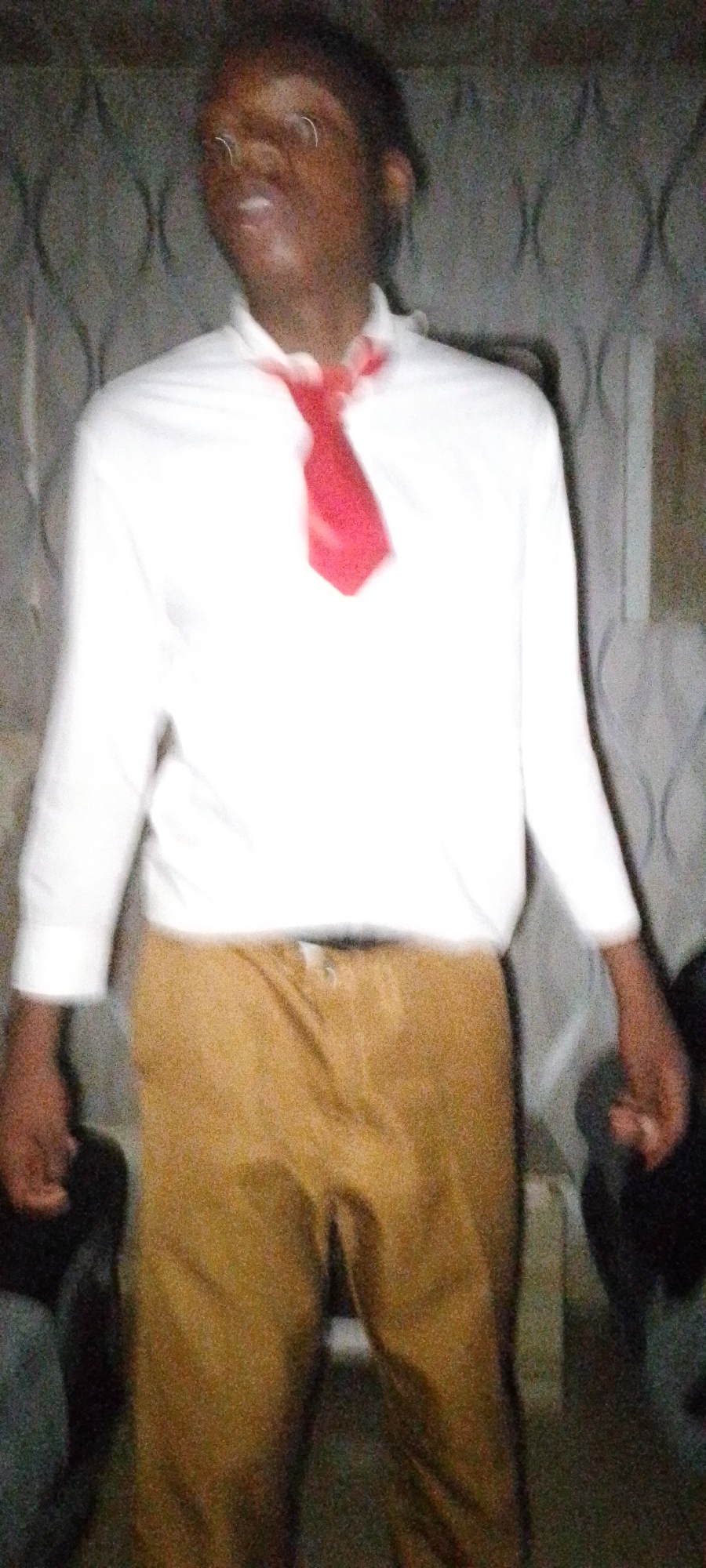 A person is standing indoors. They are wearing a long-sleeved white shirt, a red tie, and tan pants. The background is blurry, but appears to be a wall with a repeating decorative pattern. The location is Unknown, Unknown.