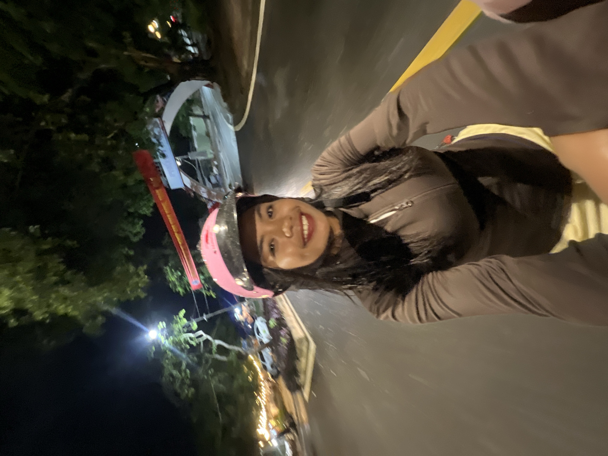A young woman wearing a pink helmet and a brown zip-up jacket is riding a motorbike at night. She is smiling at the camera, which is mounted on the front of the motorbike. The road is wet and reflects the lights from streetlamps and buildings in the background. There are trees lining the road, and in the distance, a large archway with red banners is visible. The scene appears to be in an urban or suburban area, possibly Minh Xuân Ward, Vietnam, given the mention of the location. The overall mood is lively and energetic, with the woman's smile and the bright lights creating a festive atmosphere.