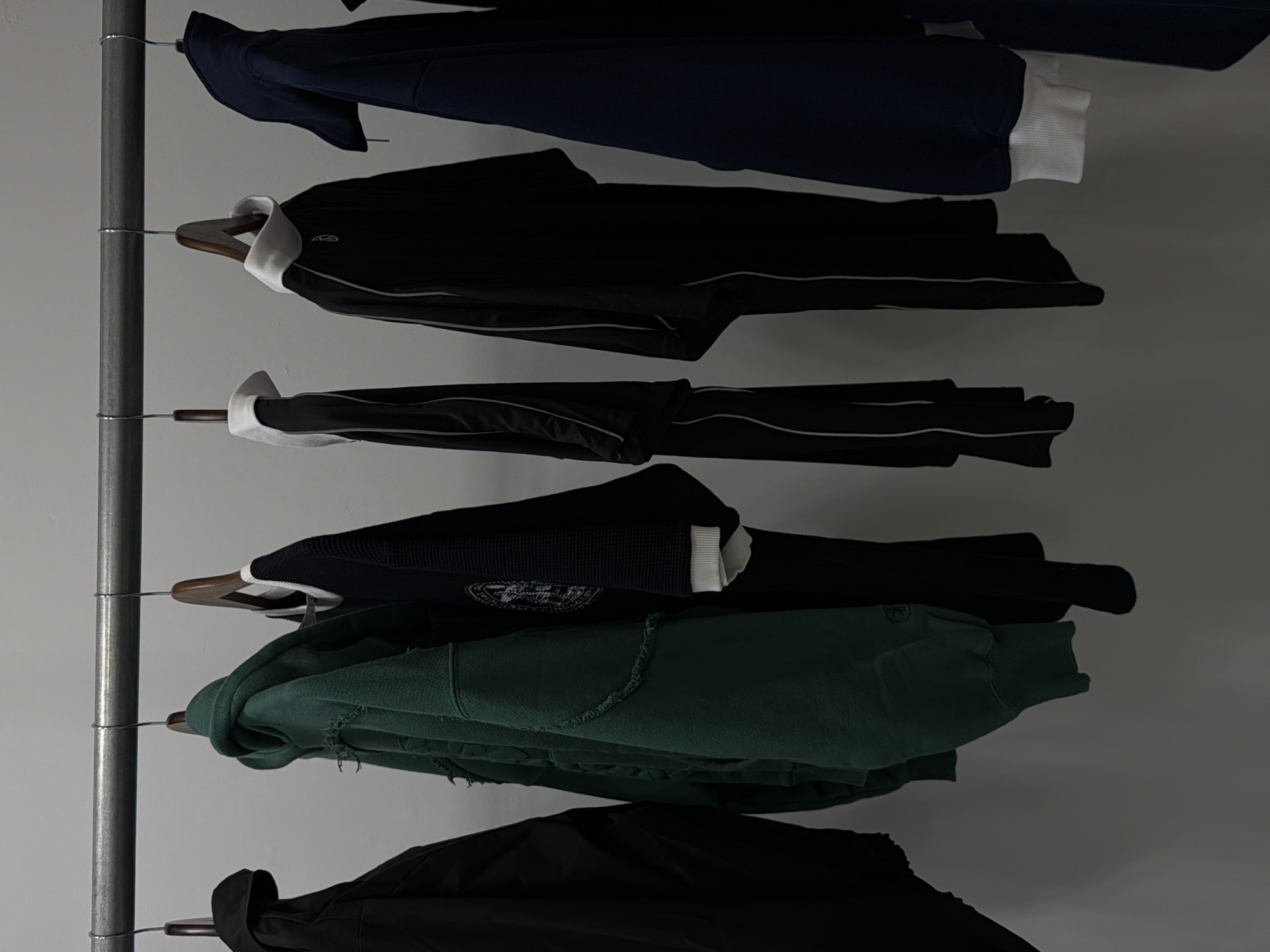 The image shows a garment rack against a grey wall. The rack is made of metal and has horizontal bars on which clothes are hung. From top to bottom, the clothing items are: a navy blue sweatshirt with white cuffs, a black jacket with white accents, a black jacket with white accents, a black knit sweater with a white cuff, a green hooded sweatshirt, and a black jacket. The background is a plain grey wall. The location is likely within New York City, United States of America, given the prompt's request, but there are no specific visual clues to confirm the city.