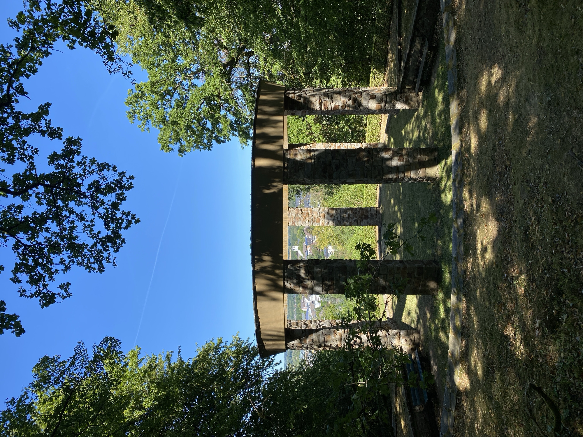 The image shows a circular stone structure with a flat roof, supported by thick stone pillars. The structure is open on all sides, offering a view of a town in the distance with buildings and trees. The ground within and around the structure is covered with grass and fallen leaves, with dappled sunlight casting shadows. Two stone benches are visible, one on each side of the structure. The scene is framed by lush green trees on either side and overhead, with a clear blue sky above. The location is the "Koppelsberg" in Altenkirchen, Germany, a scenic viewpoint overlooking the town. There are no people or animals visible in the image.