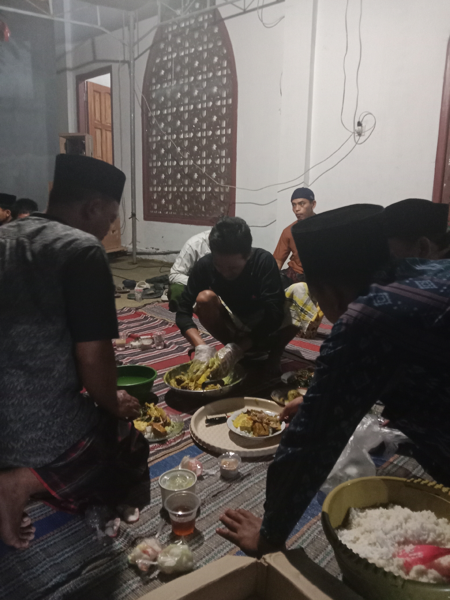 The image shows a gathering of men sitting on mats on the floor, seemingly for a meal. Several individuals are wearing traditional Muslim skullcaps, known as peci. They are gathered around various dishes of food, including what appears to be rice, curries, and other savory items. Some individuals are actively serving or preparing food, indicated by hands in bowls and the presence of serving utensils. The environment appears to be indoors, possibly in a community hall or a home, with simple walls and a decorative screen or window visible in the background. The lighting suggests it is evening or dimly lit. The presence of multiple people sharing a meal suggests a communal event or religious gathering. The image does not provide specific visual cues to pinpoint the exact location within Brangsong, Indonesia.