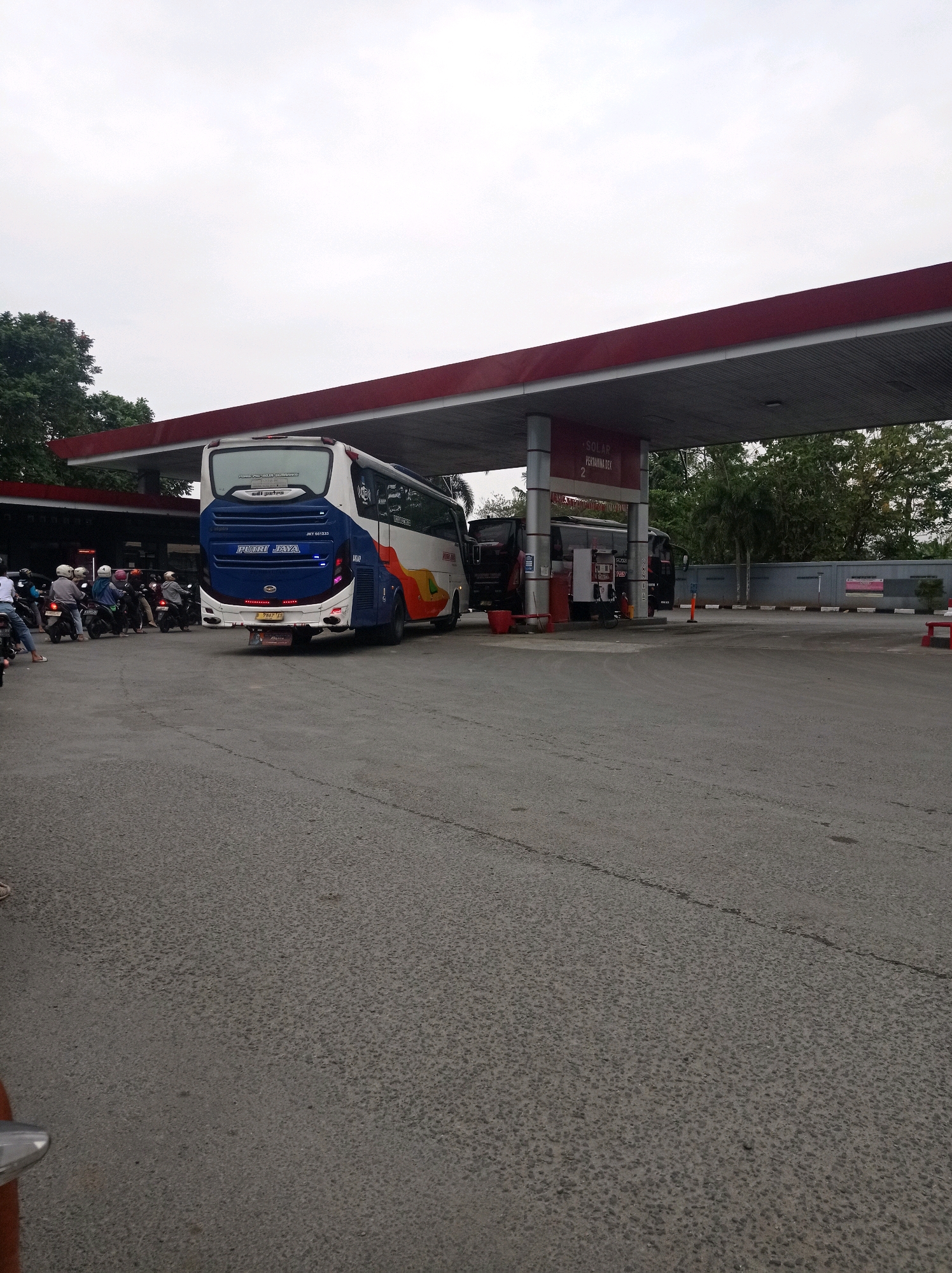 The image depicts a gas station in Bojong, Indonesia, on an overcast day. A large white and blue bus, identified as "PUTRI JAYA," is parked at a pump, with its rear facing the camera. Several motorcycles are lined up behind the bus, with their riders waiting. In the background, another bus is visible, partially obscured by the fueling station canopy. The fueling station has red trim and white pillars. Trees and a grey wall are visible behind the station. The ground is paved with asphalt.