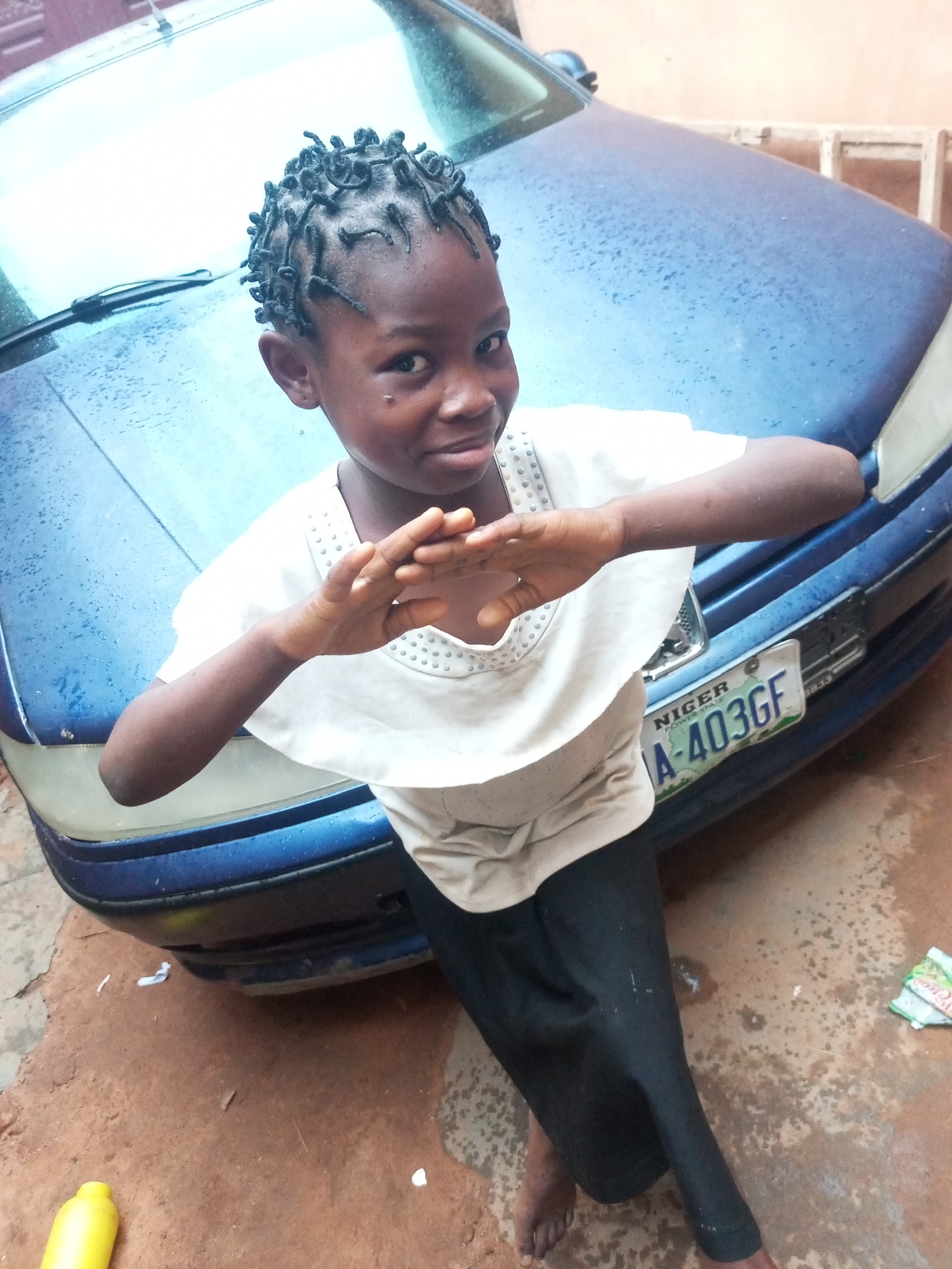 A young Black female child with braided hair and a white shirt stands in front of a blue car. She is looking at the camera and has her hands clasped in front of her chest. The car has a license plate that reads "A-403GF NIGER". The ground is made of dirt and there is some scattered debris. The overall scene appears to be outdoors in a residential area. The context of Tumfure, Nigeria, suggests a likely hot and dry climate, possibly in a rural or semi-urban setting. The blue car may be a common mode of transportation in this region. The child's attire, a white shirt and dark pants, are practical for warm weather. The lighting suggests it is daytime.