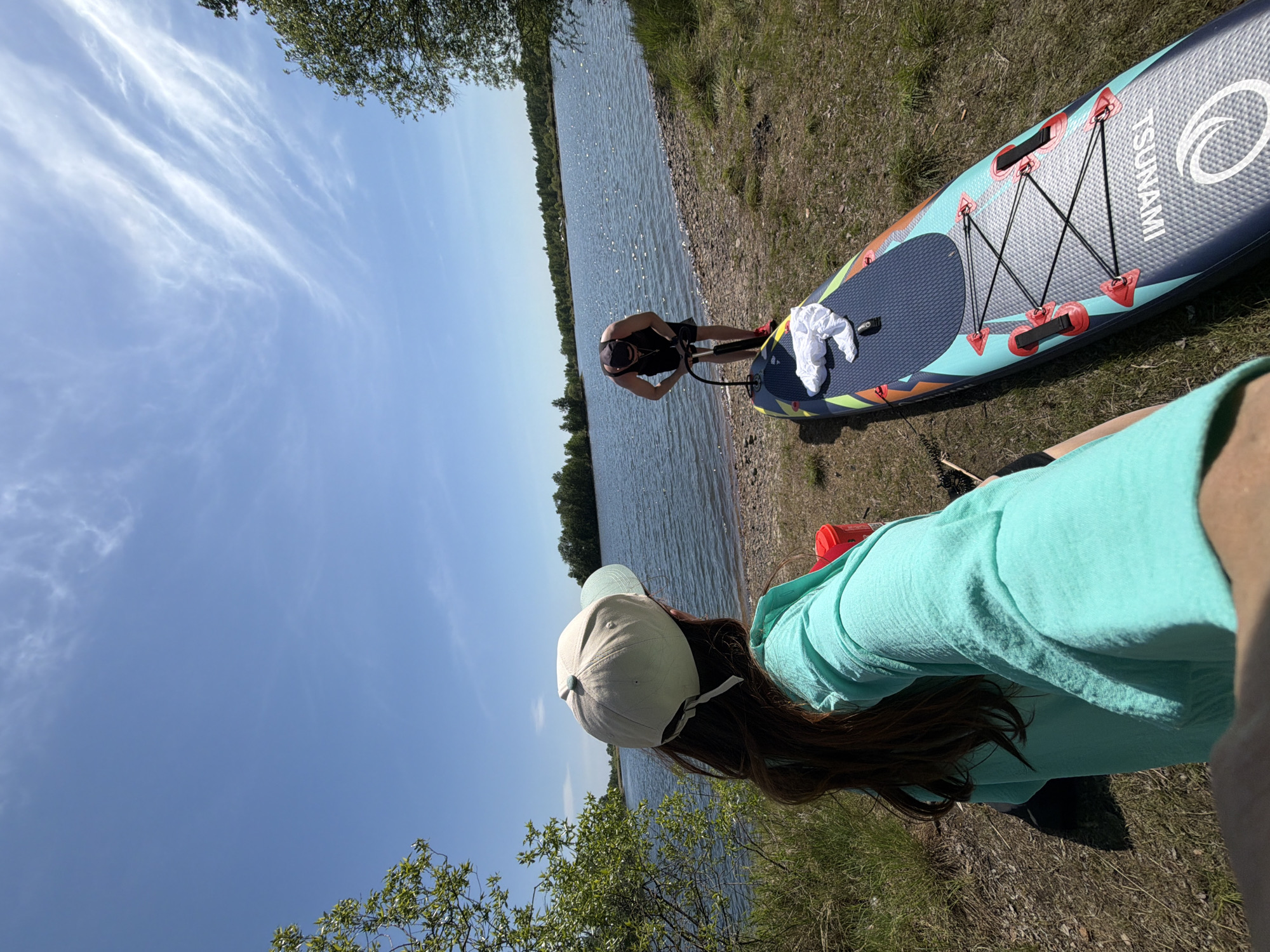 On a bright day with a blue sky and wispy clouds, two individuals are present near a body of water, likely a lake or river, in Birobidzhan, Russia. The foreground features a person wearing a light turquoise shirt and a light-colored baseball cap, positioned to the right of the frame and facing the camera. Their long brown hair is visible under the cap. Adjacent to this person, a deflated, blue and turquoise paddleboard lies on the grassy bank. A white cloth rests on the board. Further into the scene, another person stands near the water's edge, appearing to inflate the paddleboard using a manual air pump. This person is wearing a dark-colored top and shorts. The water is calm with a slightly reflective surface, and a line of trees is visible on the opposite bank. The ground where the paddleboard and people are situated is covered with grass and small pebbles.